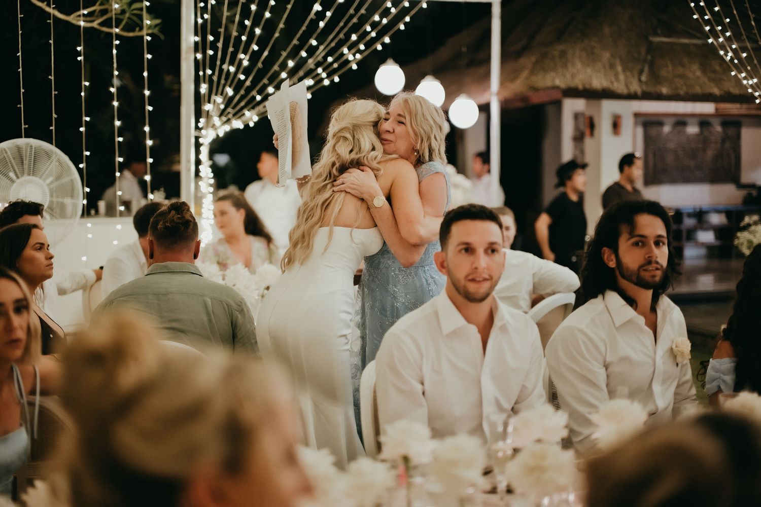 Bride embraces a guest warmly at a romantic evening wedding reception adorned with fairy lights.