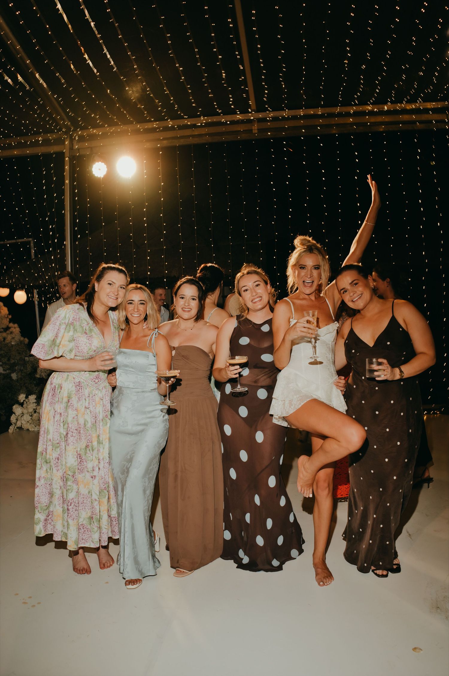 Group of six female wedding guests posing and dancing together under string lights at evening reception.