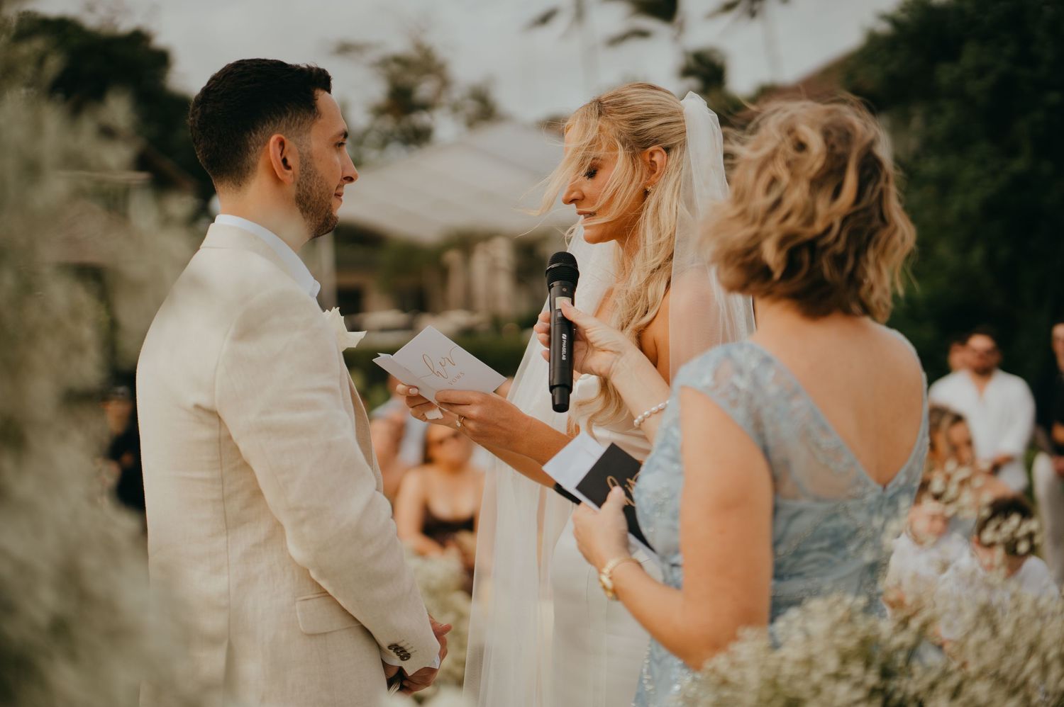 Blonde bride reads vows at outdoor ceremony while groom in white suit listens attentively.