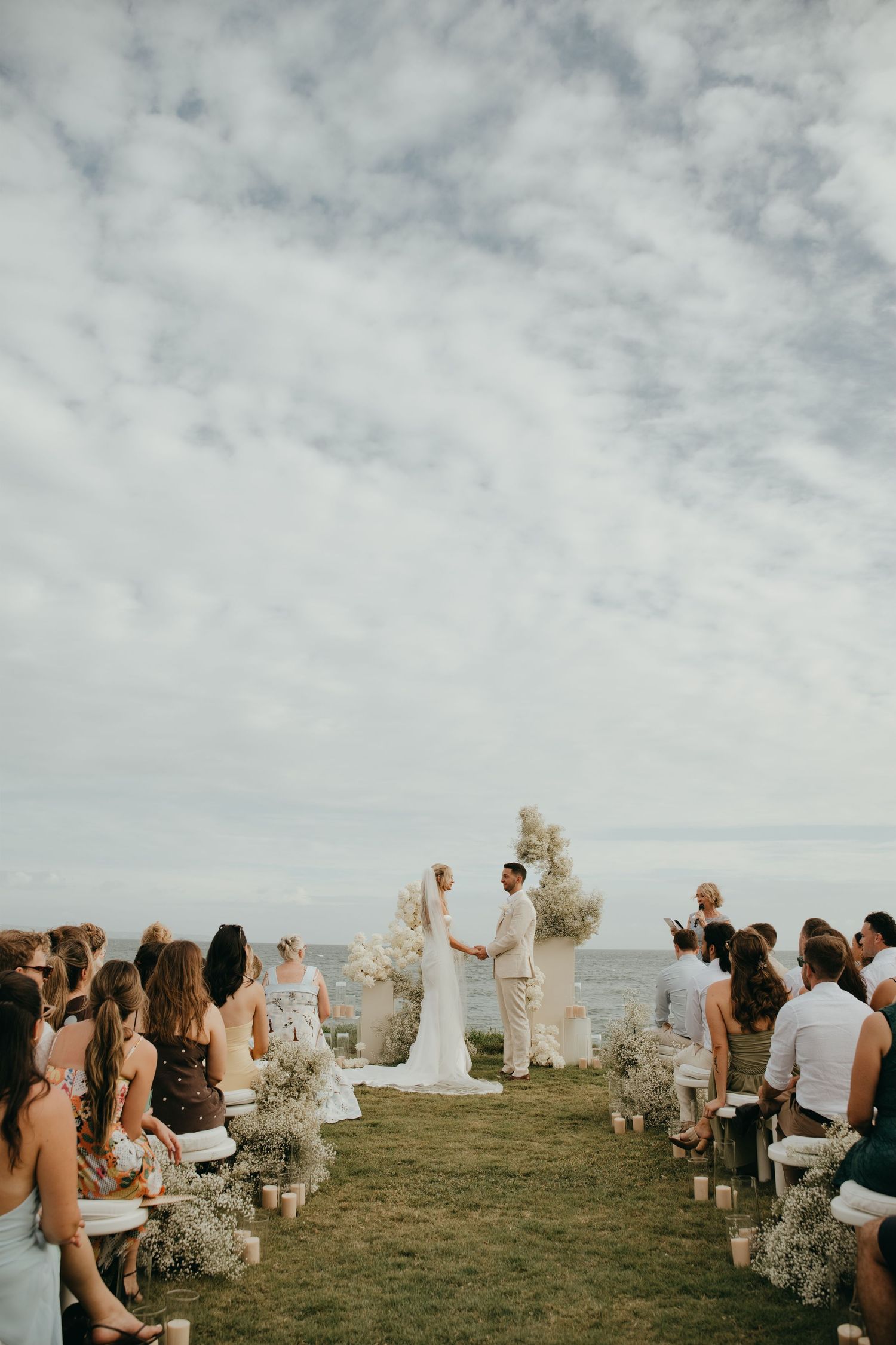 Couple exchanges vows at a cliffside ocean wedding ceremony with white floral arch and seated guests.