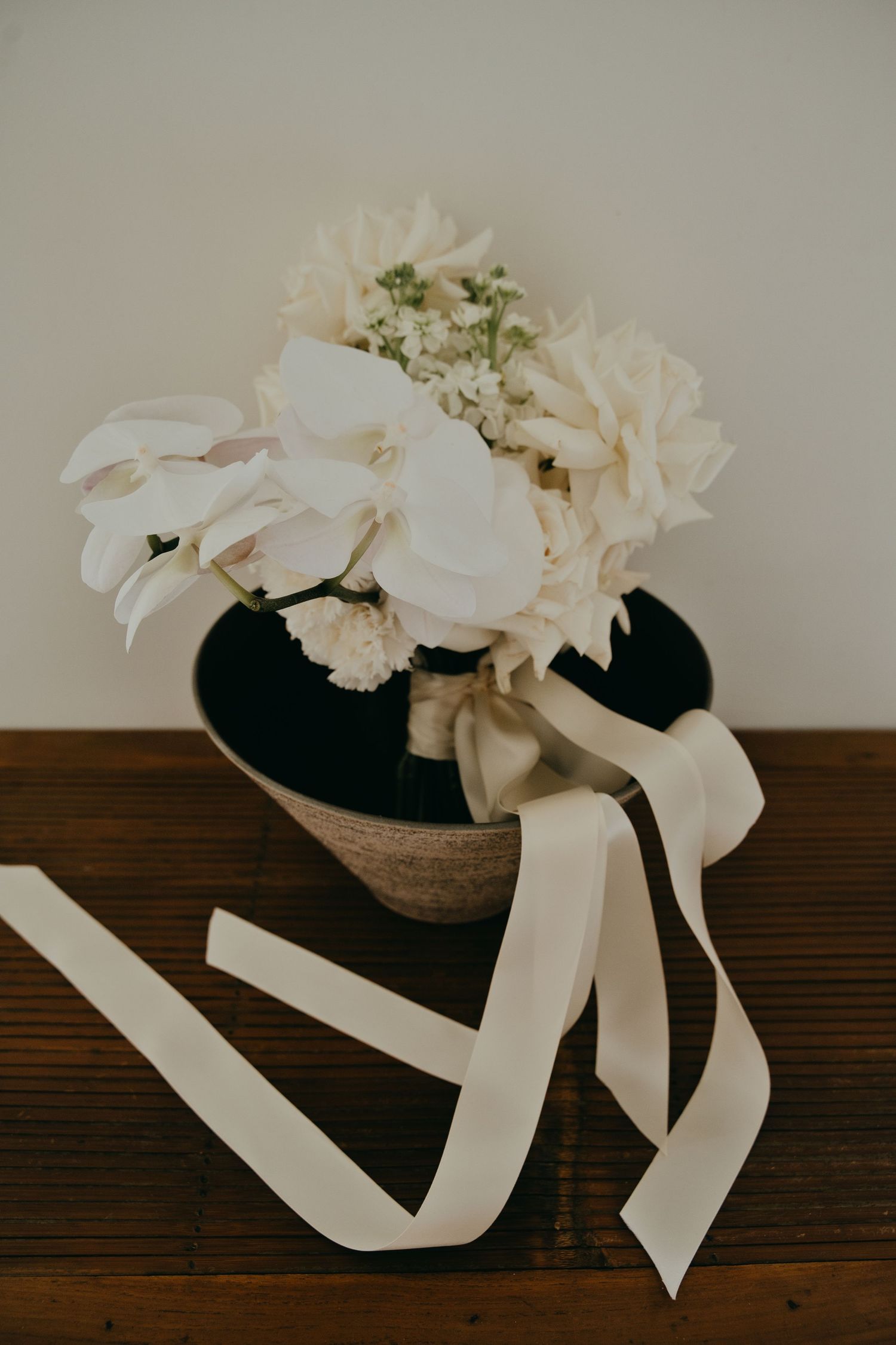 Elegant white bridal bouquet with orchids and hydrangeas in a vase with flowing ivory ribbon on wood table.