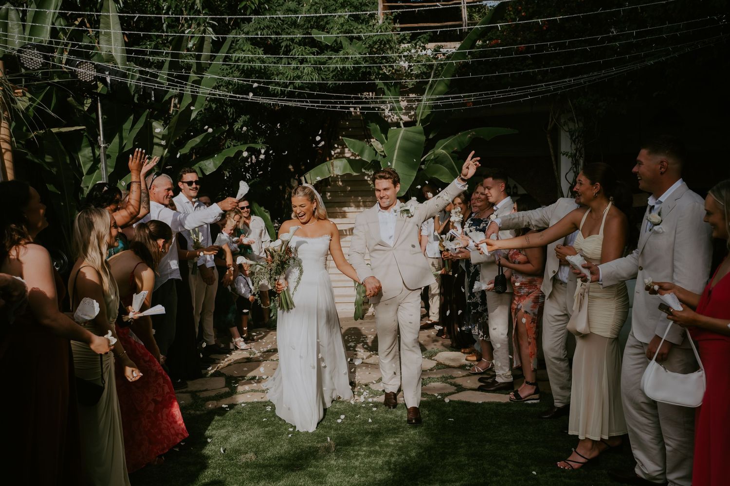 Newlyweds celebrate joyful confetti exit surrounded by cheering guests at tropical outdoor wedding.