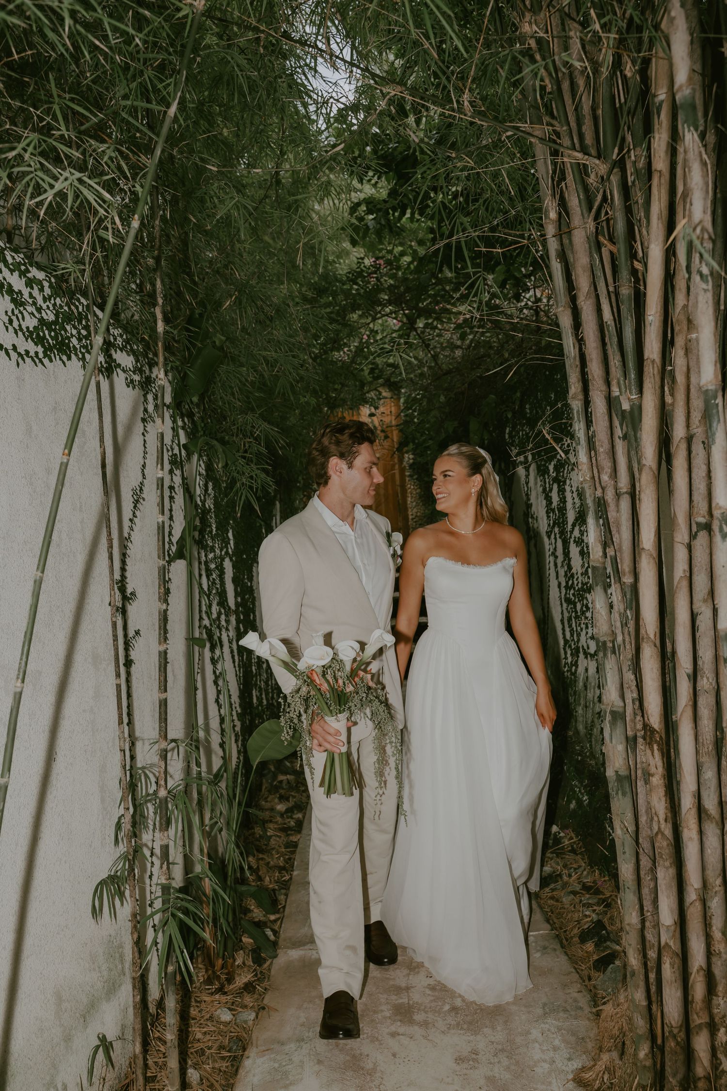 Bride and groom walking hand in hand through lush bamboo garden pathway on wedding day.