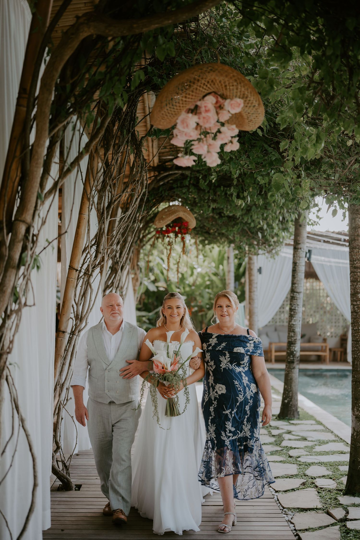 Bride walks down outdoor aisle with parents at lush tropical garden wedding.