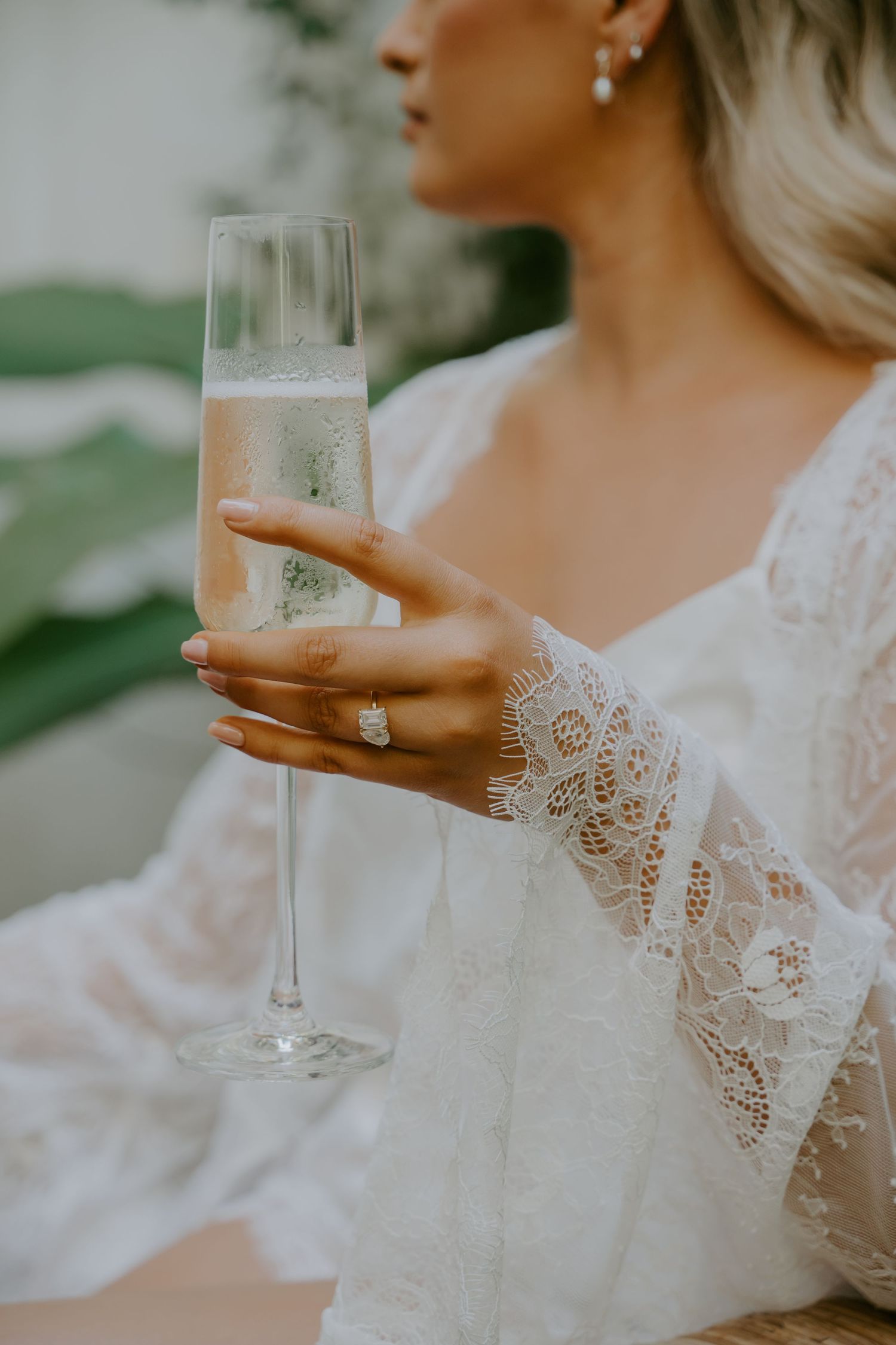 Bride in lace robe holding champagne flute with diamond engagement ring during wedding morning preparations.