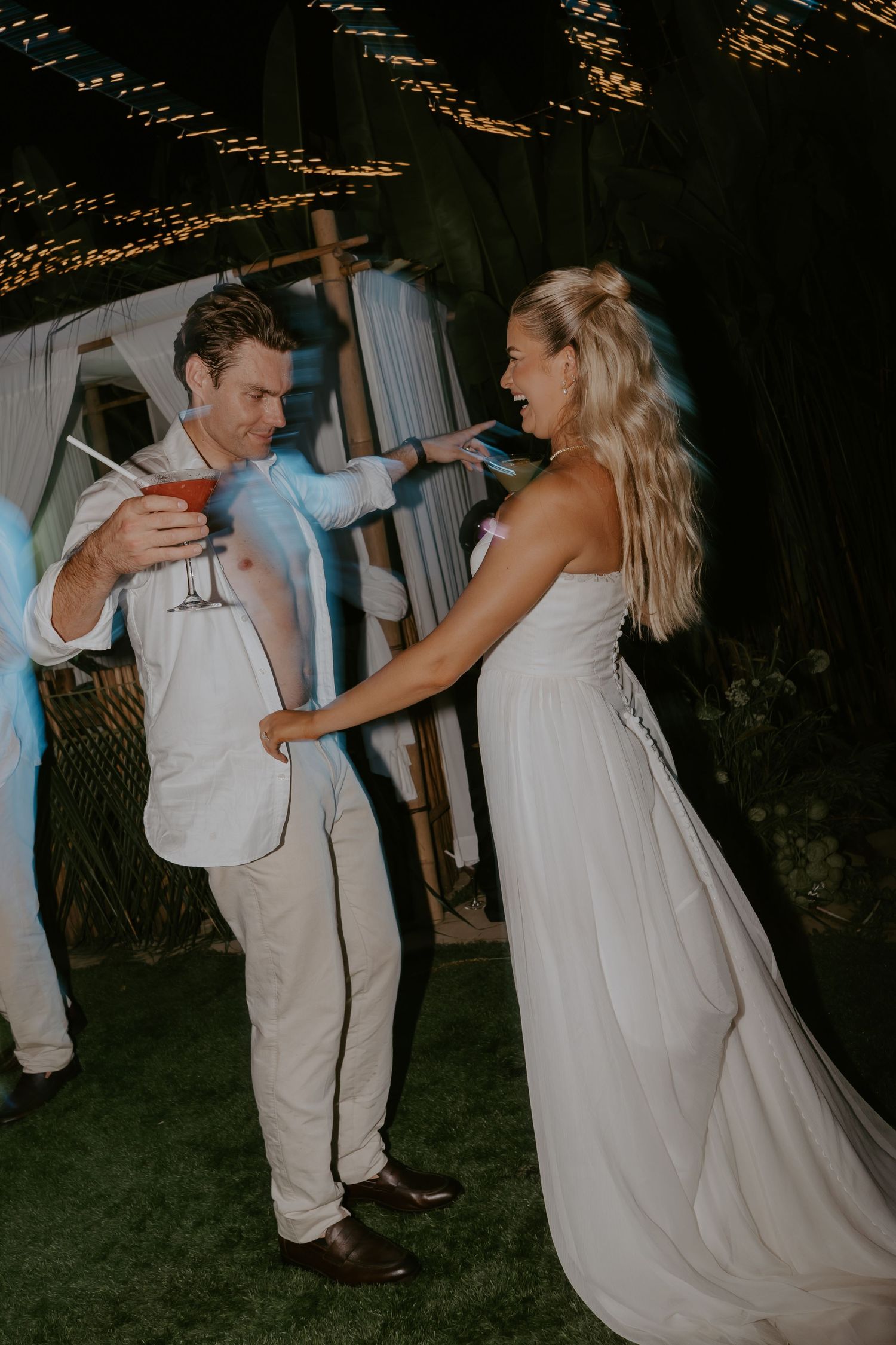 Bride and groom laugh together outdoors at night, groom holding a red cocktail at wedding.