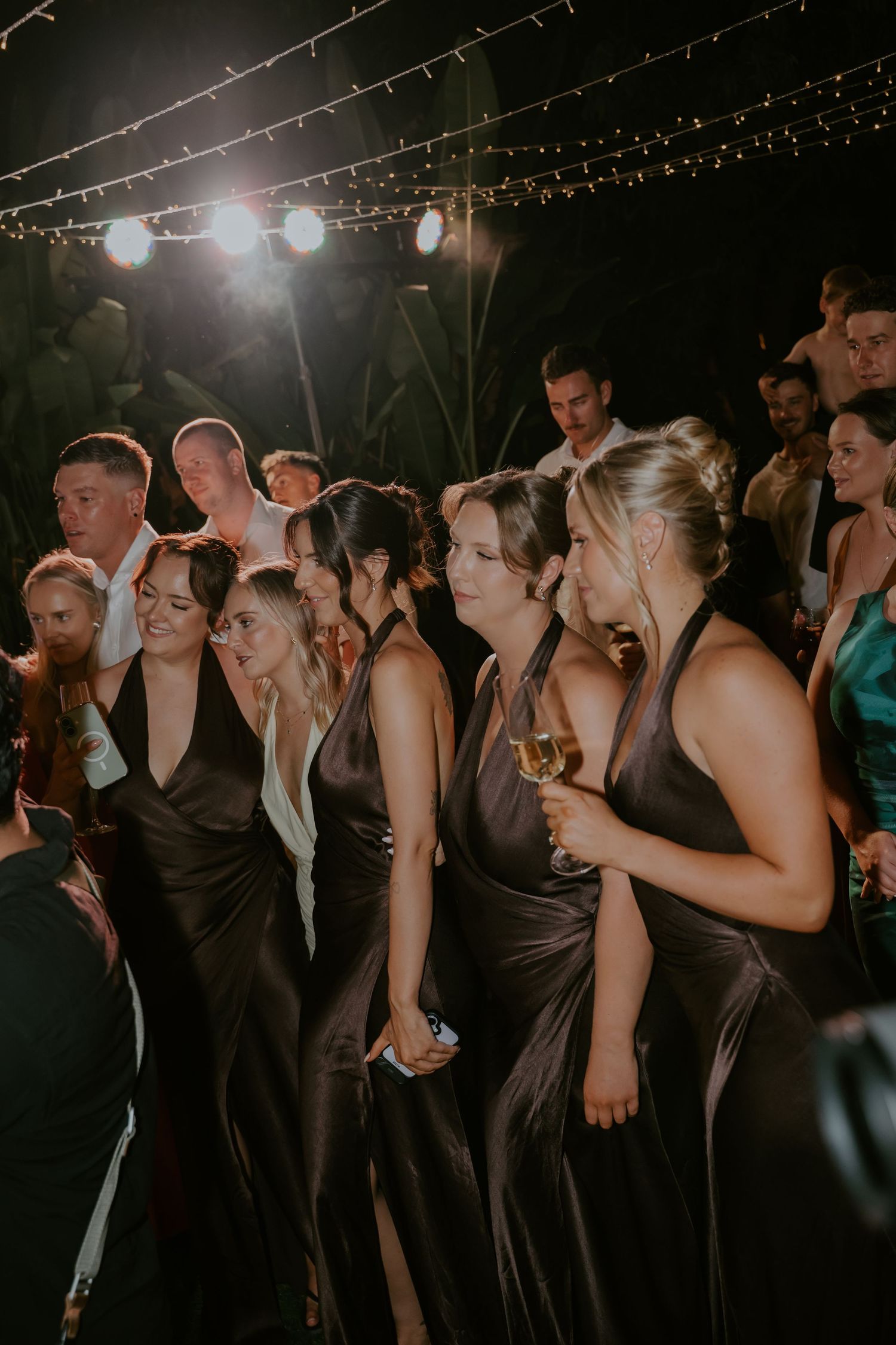 Bridesmaids in elegant dark satin gowns dancing and celebrating at an outdoor wedding reception under string lights.
