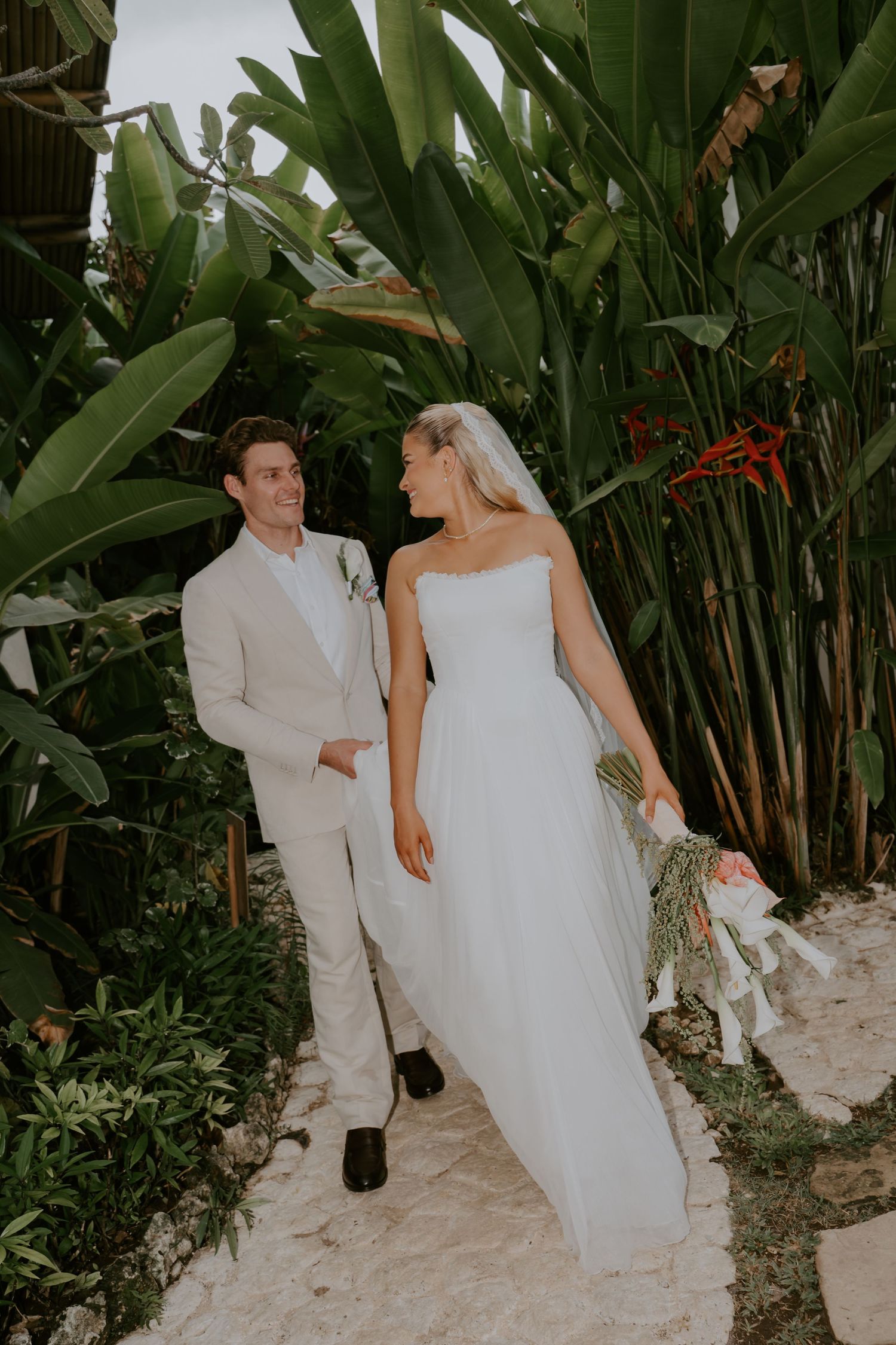 Newlyweds walk hand in hand through lush tropical garden after wedding ceremony.