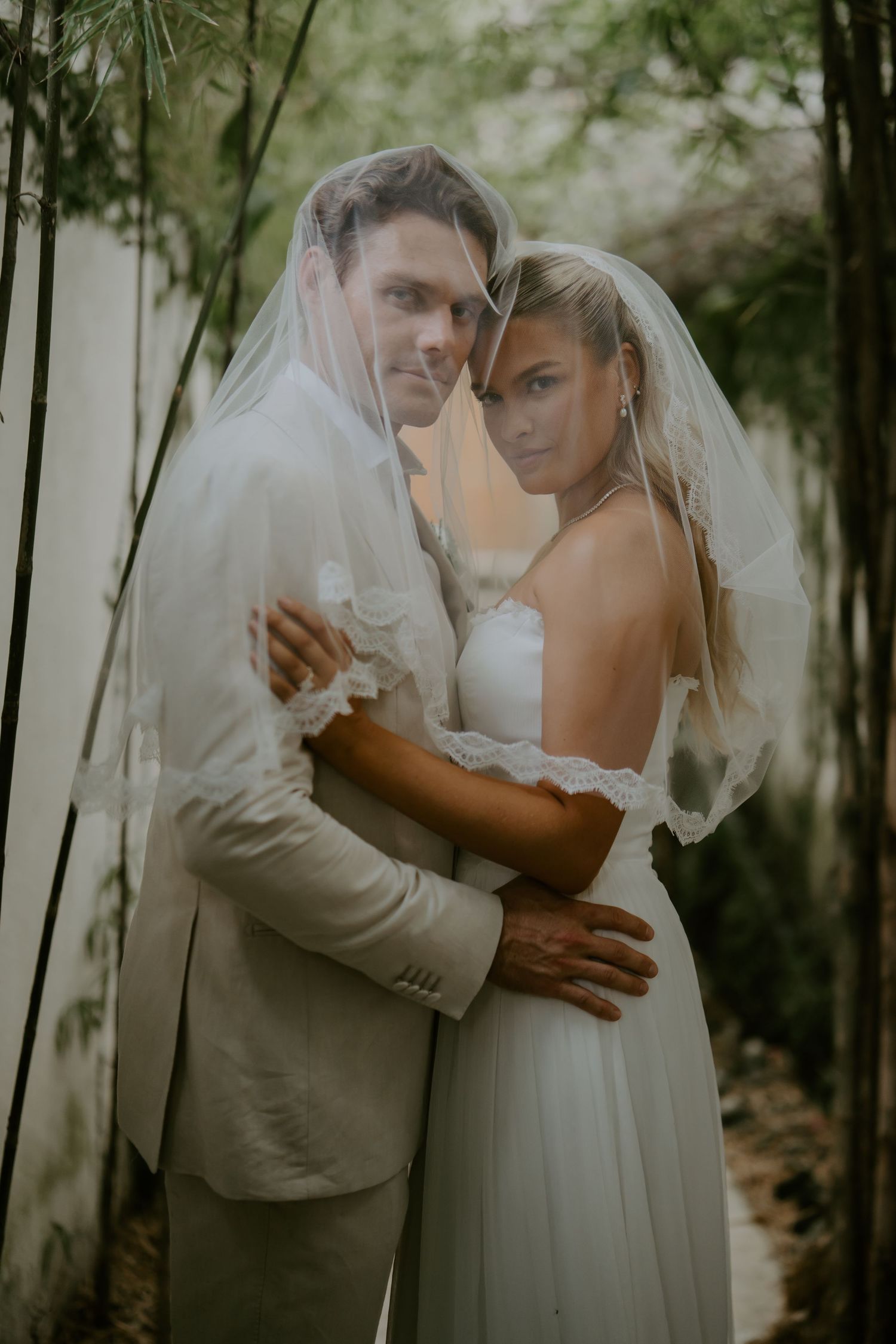 Bride and groom posing together under a flowing veil surrounded by bamboo greenery.