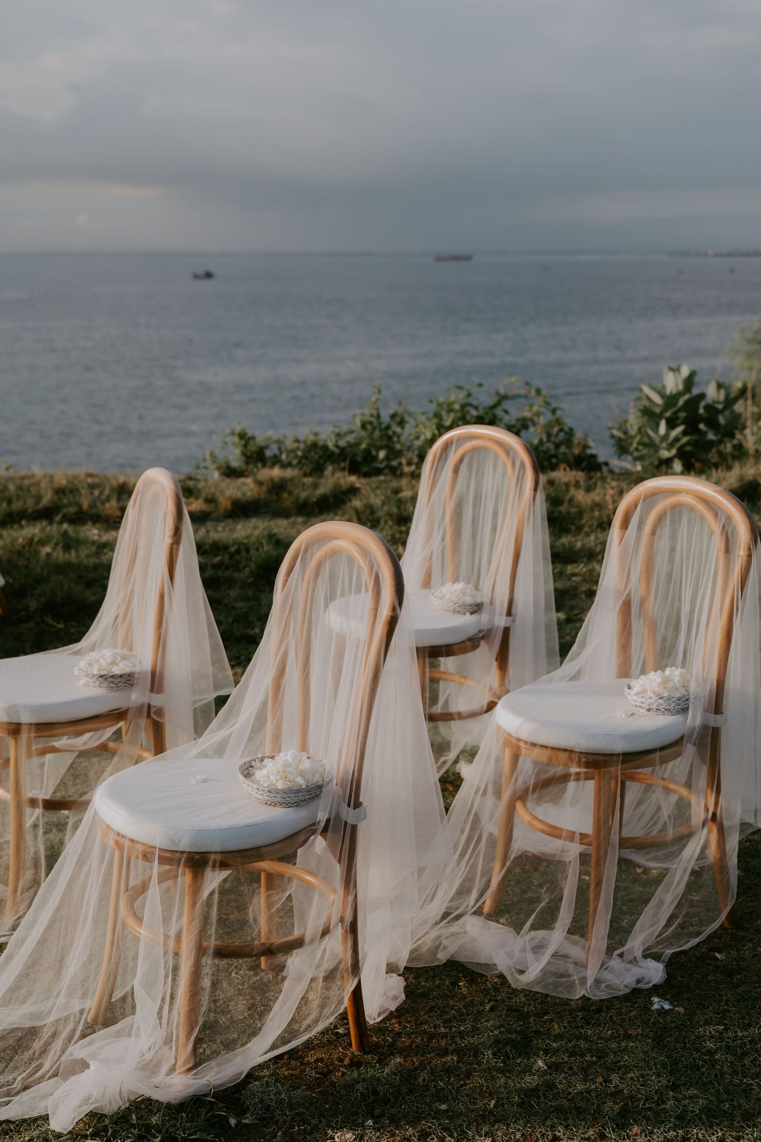 Elegant rattan chairs draped with white tulle overlooking the ocean at a seaside wedding ceremony.