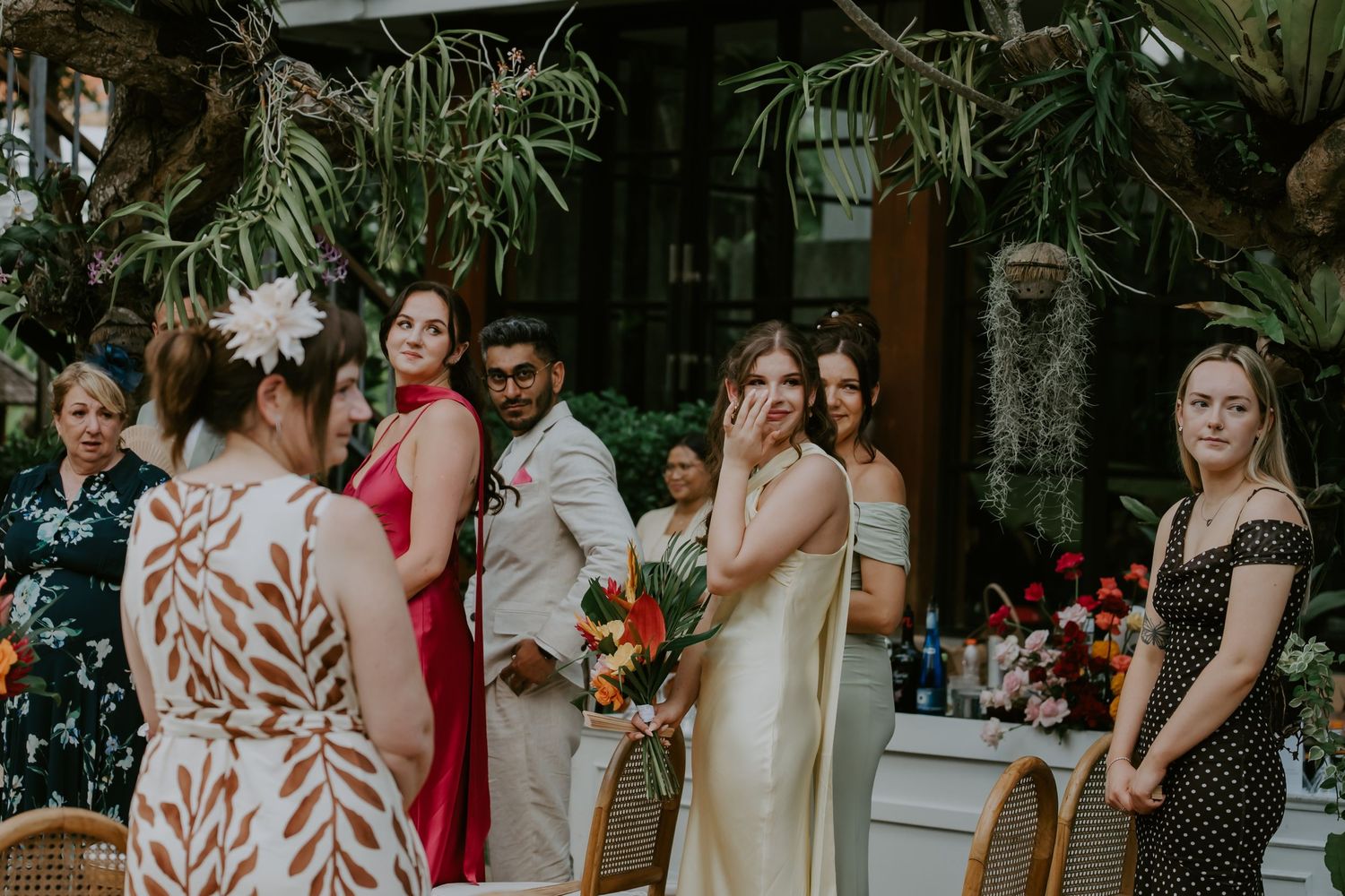 Emotional bride wiping tears during outdoor wedding ceremony surrounded by guests in colorful attire and lush tropical greenery.