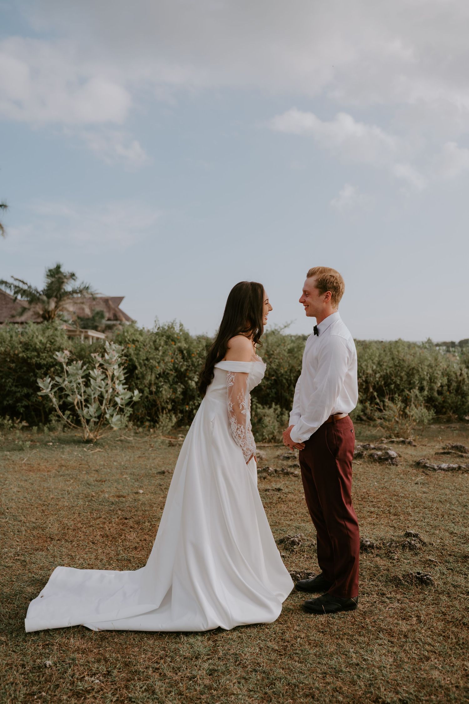Bride in off-shoulder lace wedding gown faces groom in burgundy pants outdoors.