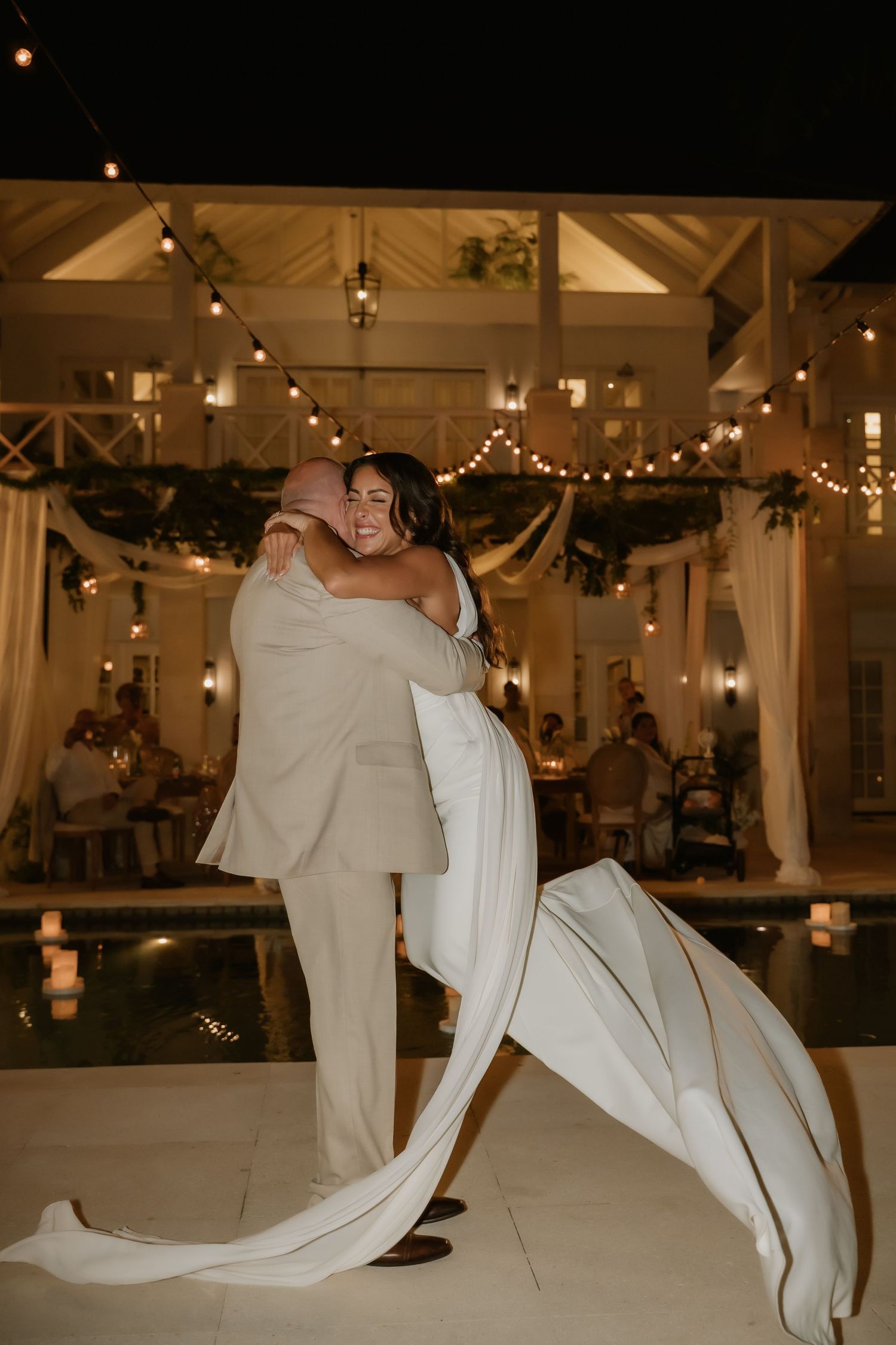 Groom lifts smiling bride in wedding dress beside candlelit pool during outdoor evening reception.