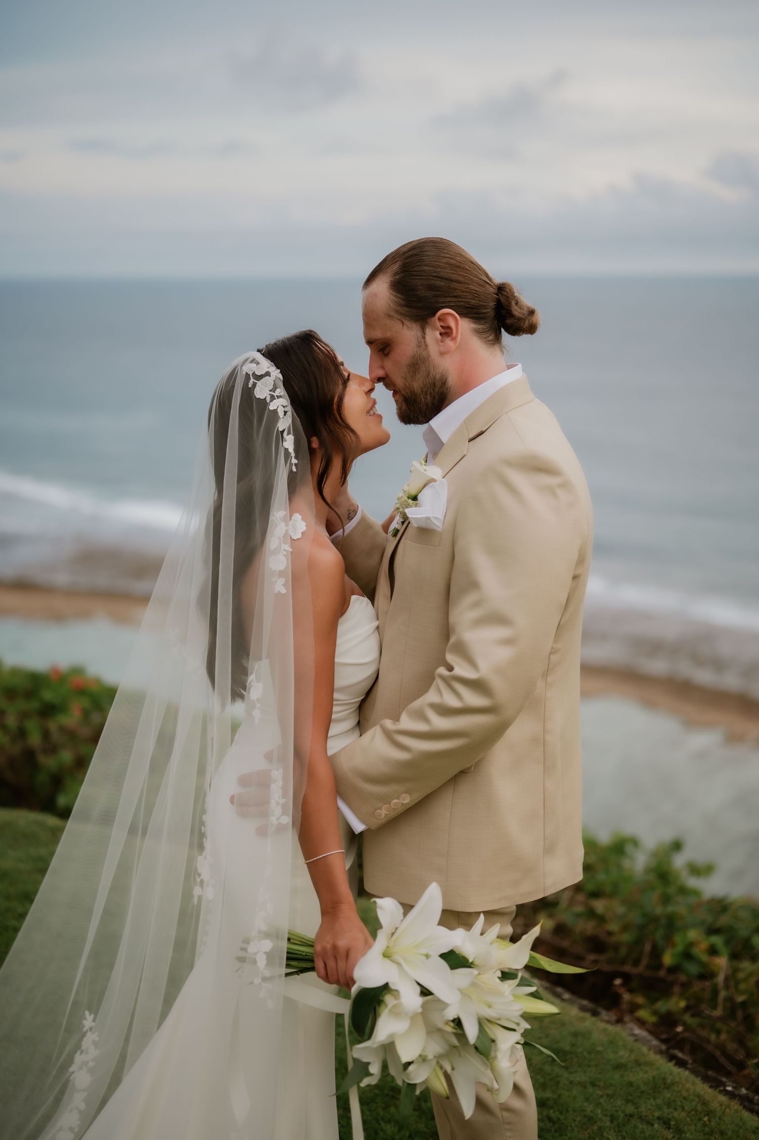 Bride and groom sharing a romantic moment with ocean backdrop, white lily bouquet and veil.