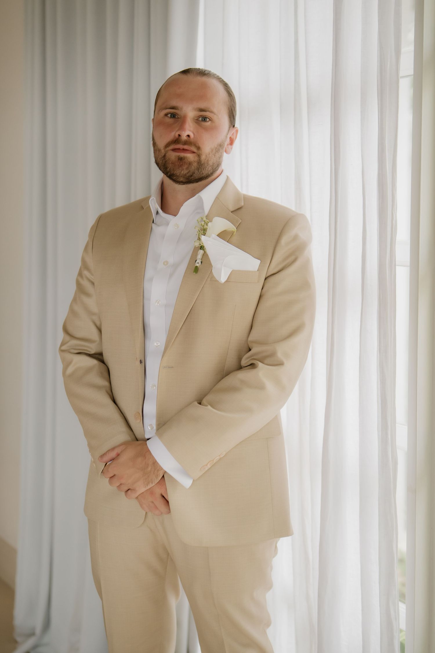 Groom in a beige suit with white boutonniere standing before sheer curtains on wedding day.