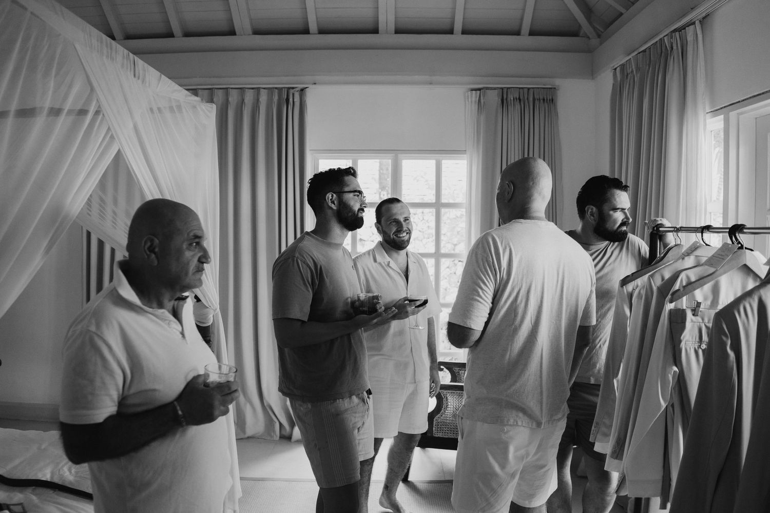 Groomsmen getting ready in a bright room, selecting suits from a rack in black and white photo.