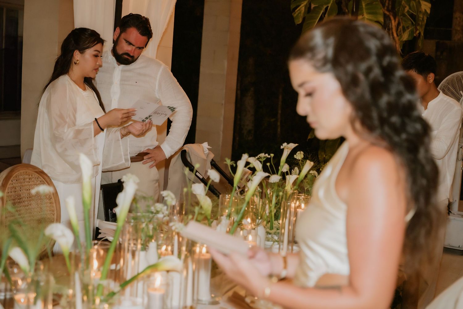 Couple in white attire lights candles at elegantly decorated wedding reception dinner table.