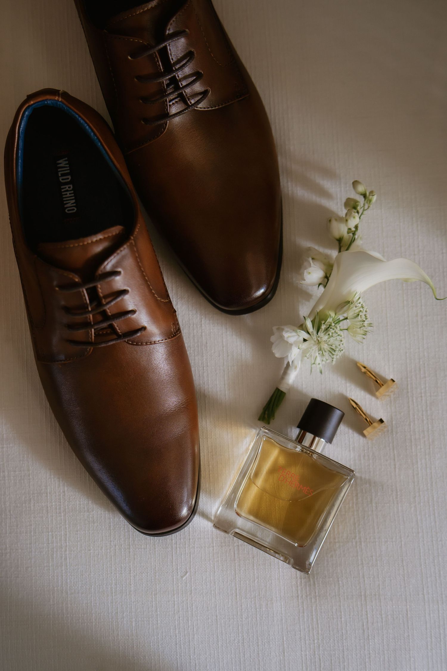 Brown leather dress shoes with cologne, white flowers, and gold cufflinks arranged on a white surface for a wedding.