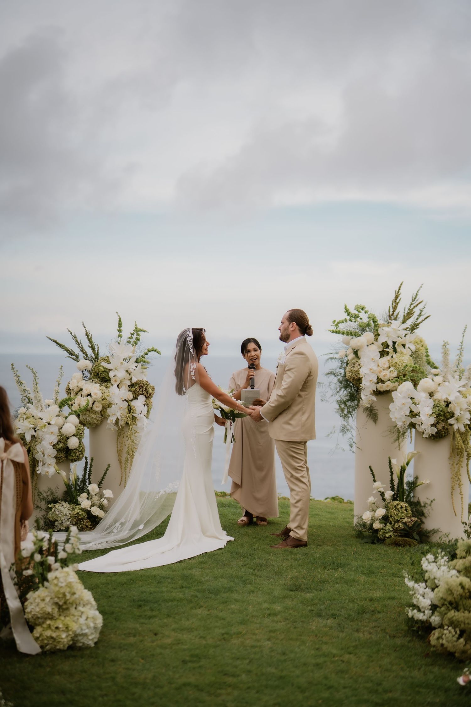 Couple exchanges vows at outdoor wedding altar adorned with white floral arrangements overlooking the ocean.
