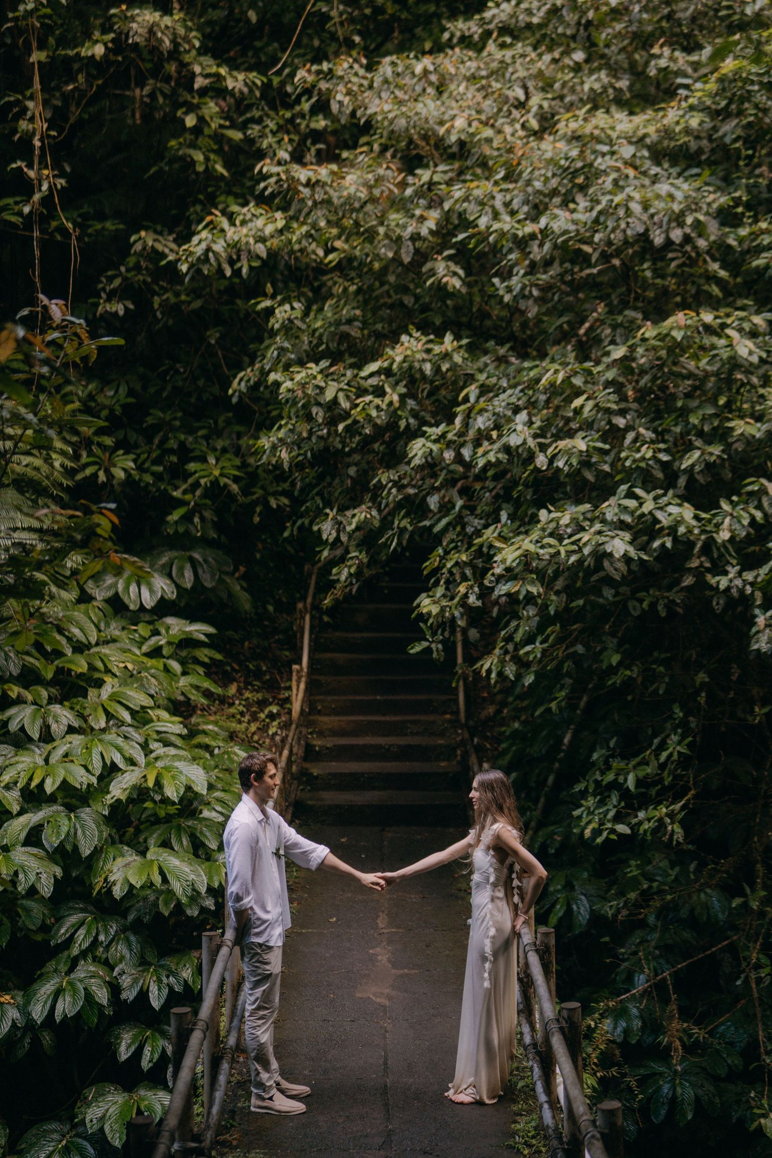 A romantic couple holds hands in a lush garden setting with green foliage creating a natural backdrop.