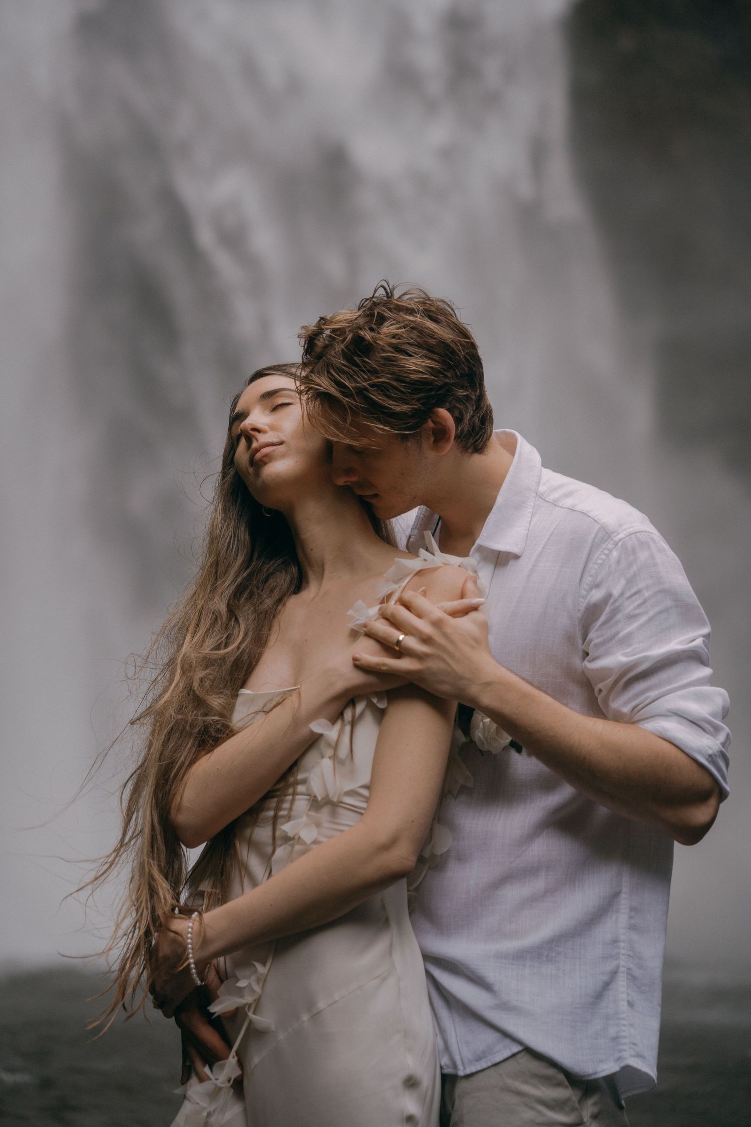 Romantic couple embraces in front of misty waterfall backdrop, creating ethereal and moody atmosphere.