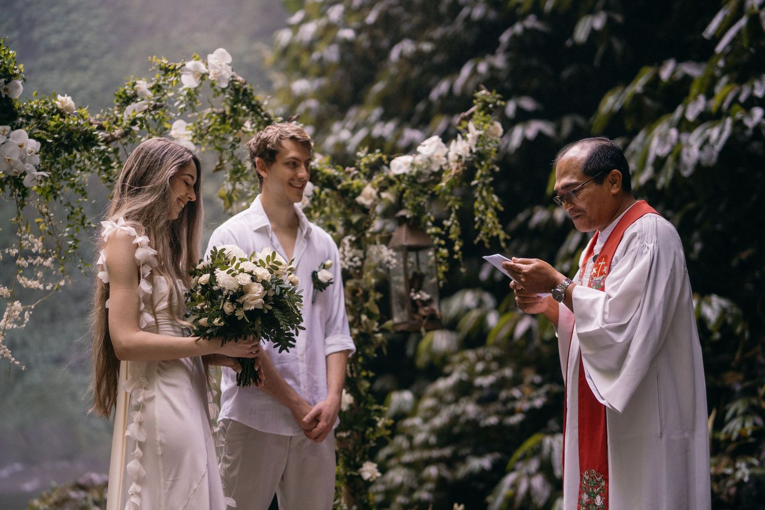 A wedding ceremony takes place in a garden with blooming white roses as an officiant reads from a book.
