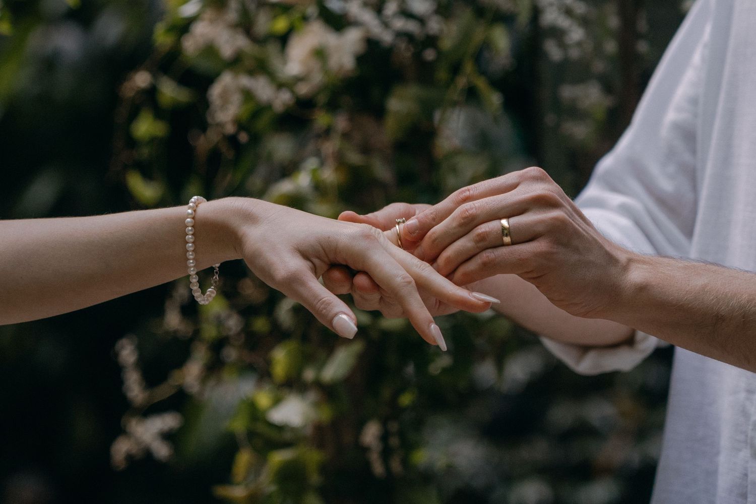 Close-up sequence of hands exchanging wedding rings during an outdoor ceremony with white flowers in background.