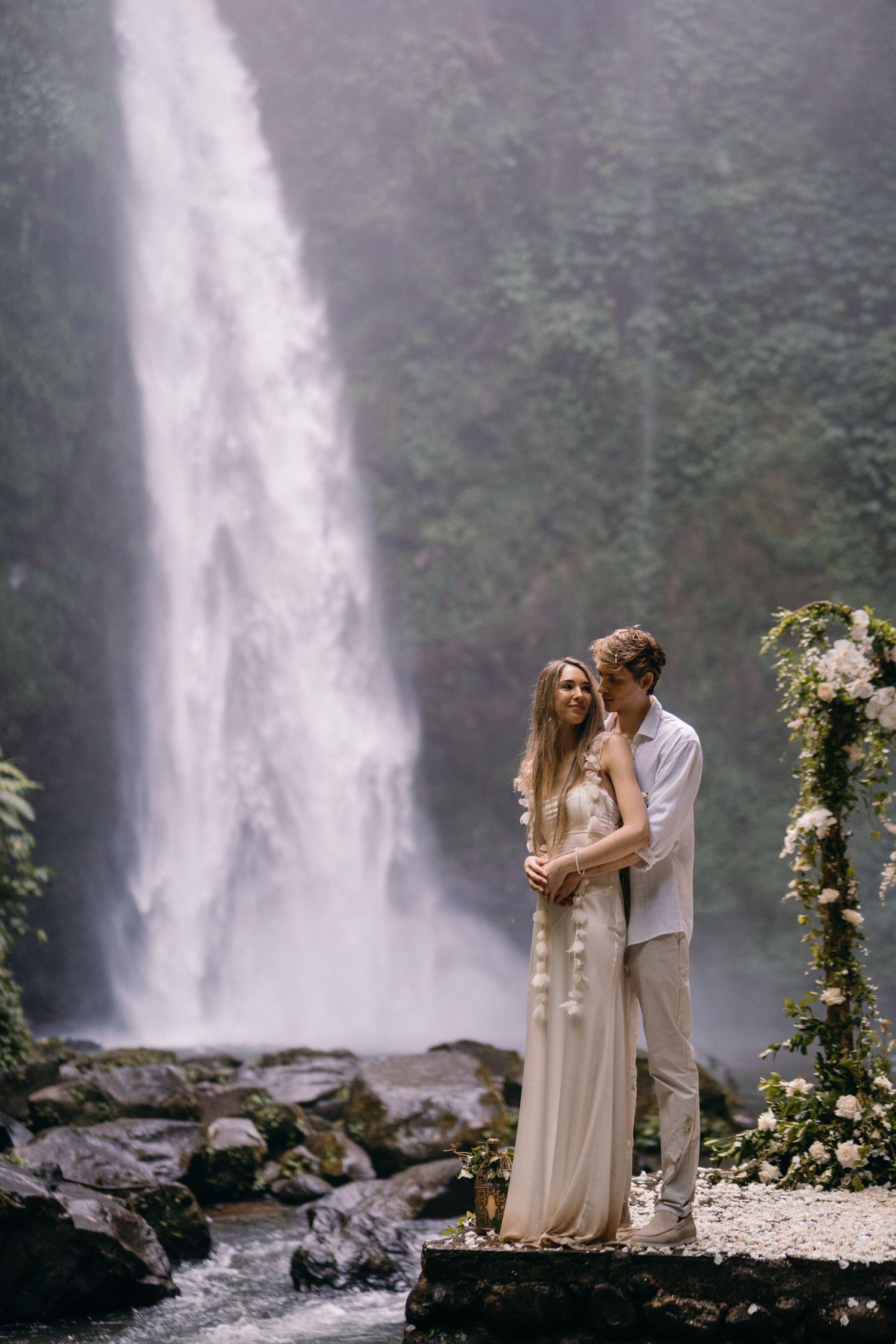 Romantic couple embraces near a majestic waterfall with floral arch decorations in a misty natural setting.