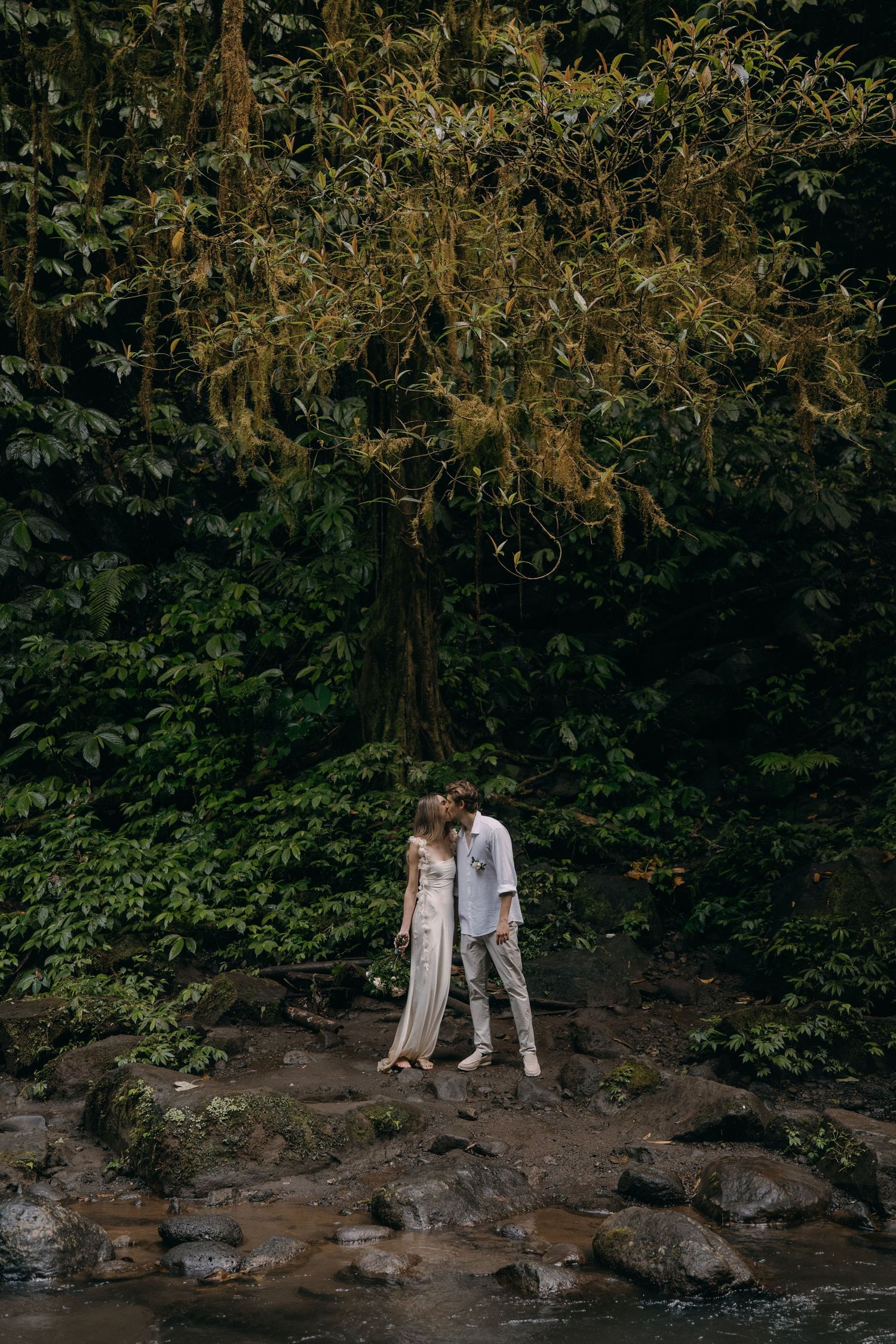 Two people in light colored clothing explore a rocky creek bed surrounded by lush greenery and overhanging trees.