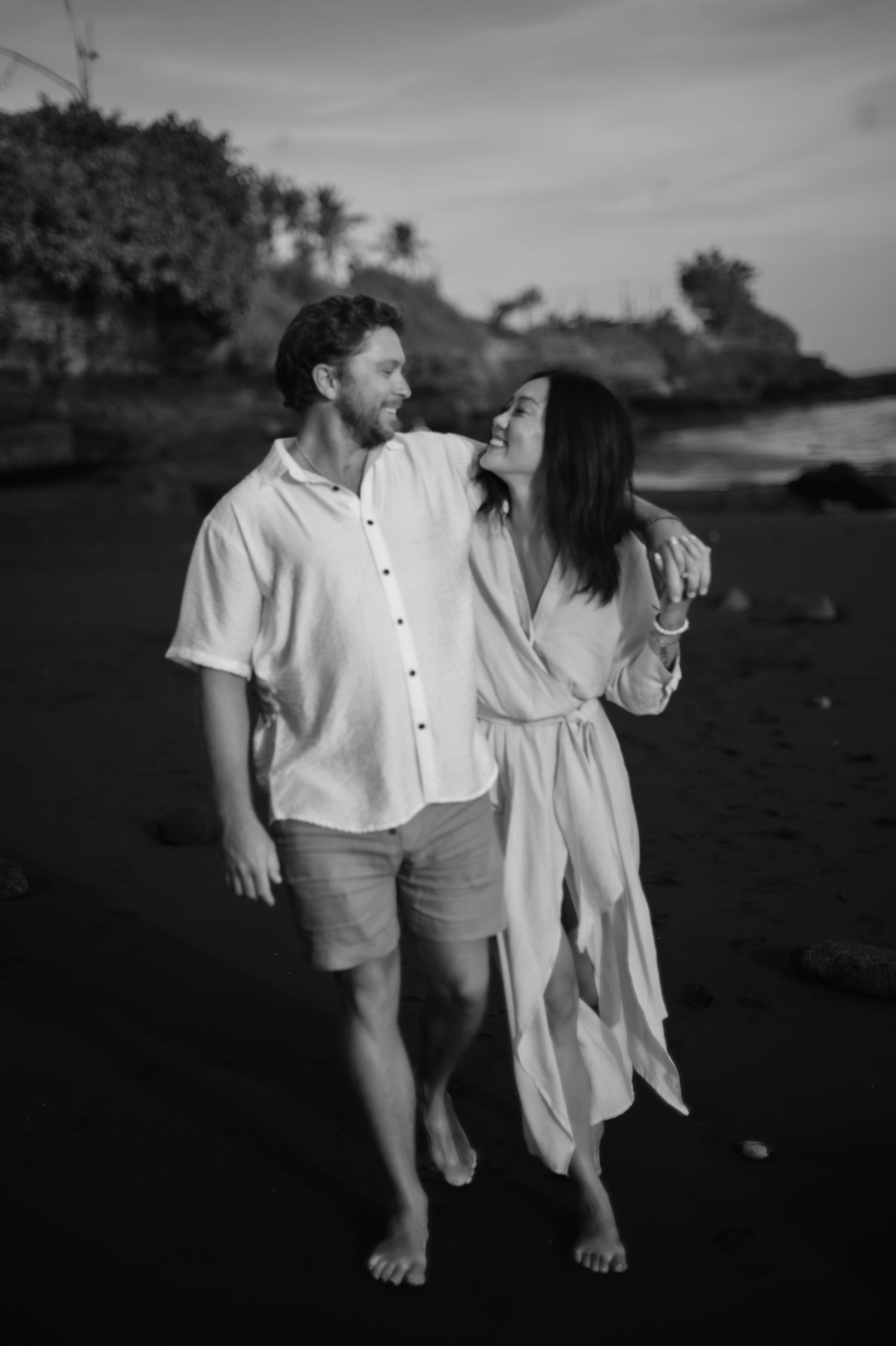 A couple in white outfits walk barefoot on a beach at sunset sharing a romantic moment in black and white photography.