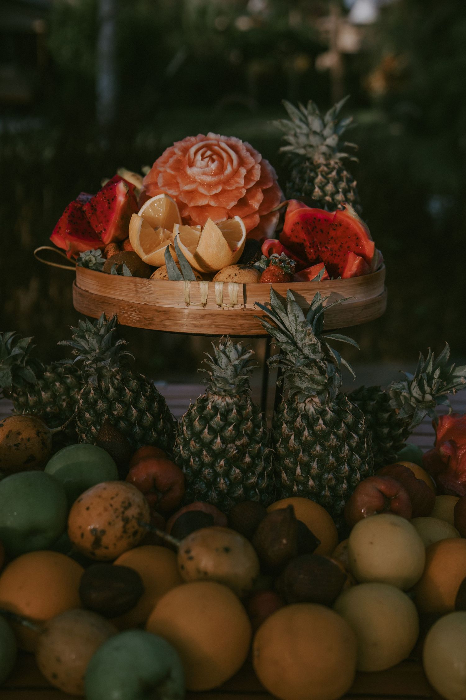 A moody still life arrangement of tropical fruits with pineapples and citrus on a wooden tiered display with carved fruit decor.