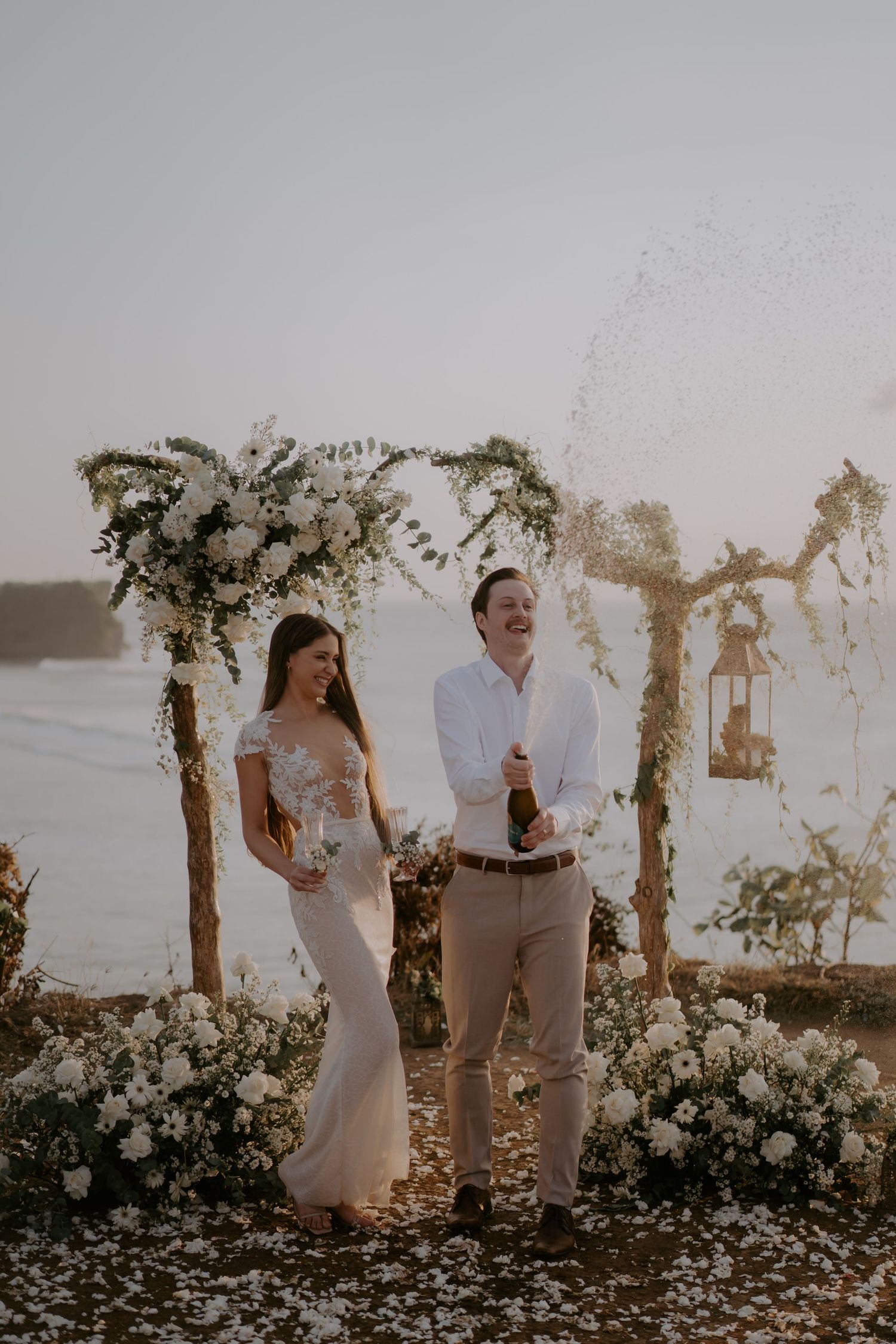 A couple in light-colored attire poses together in a garden setting with flowering trees during sunset.
