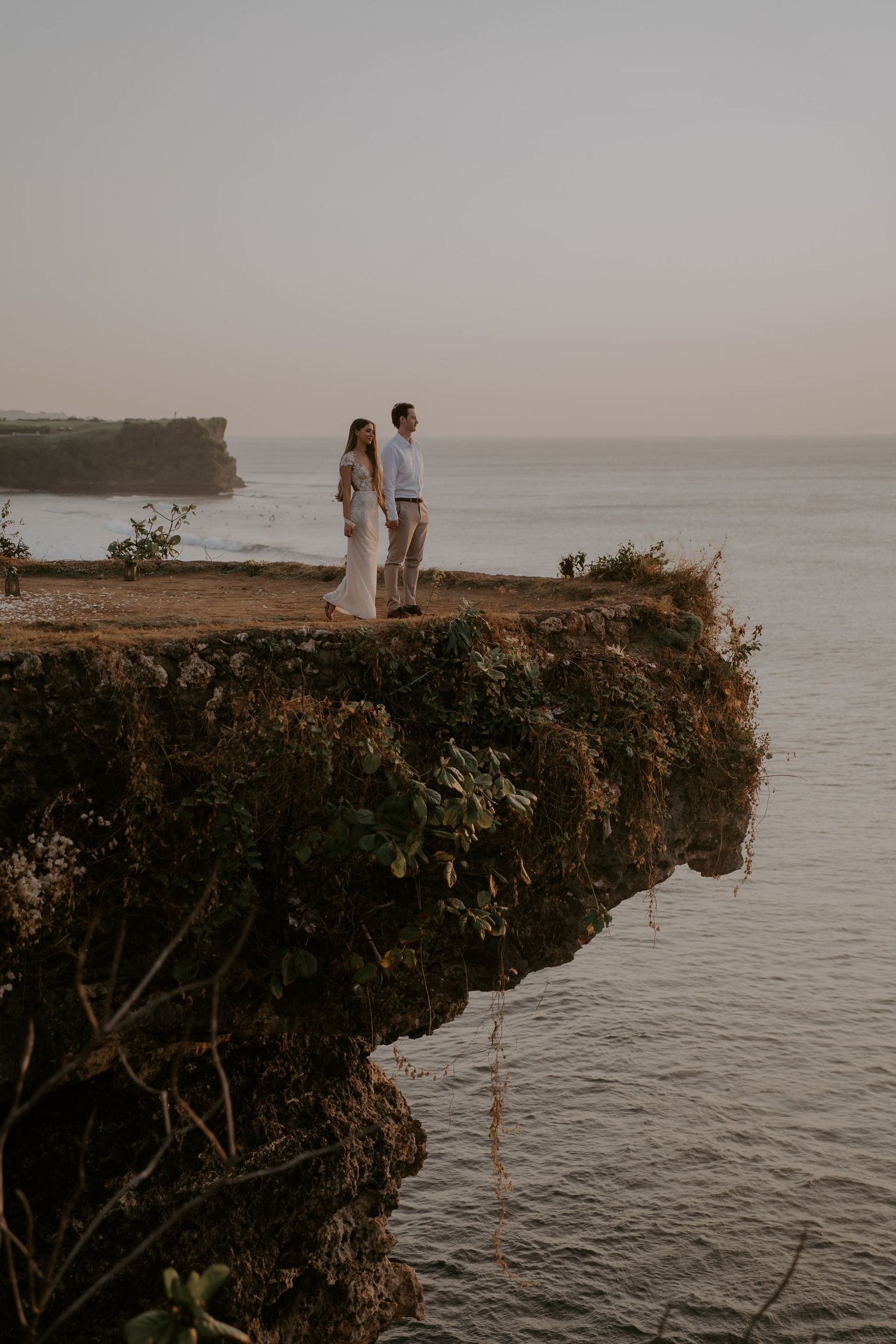 Two figures stand silhouetted on a dramatic ocean cliff edge at sunset overlooking the vast sea below.