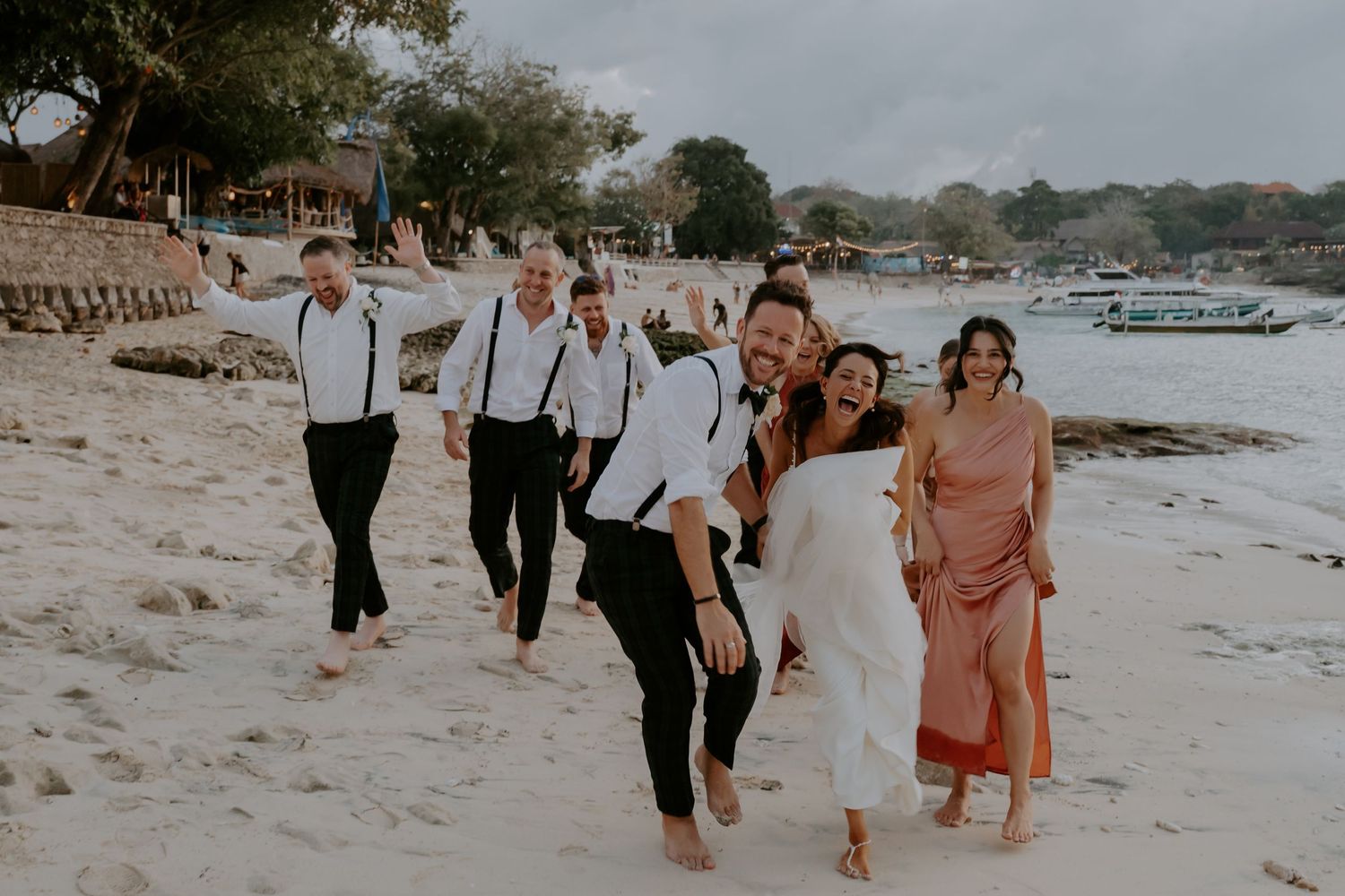 Wedding party walks along a tropical beach with palm trees and boats in the background at sunset.