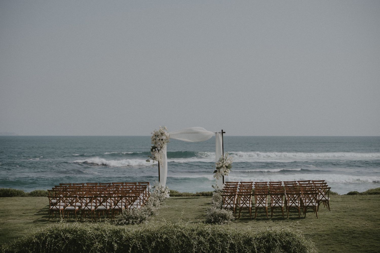 Rustic wooden chairs arranged for an outdoor beach wedding ceremony with ocean waves in the background.