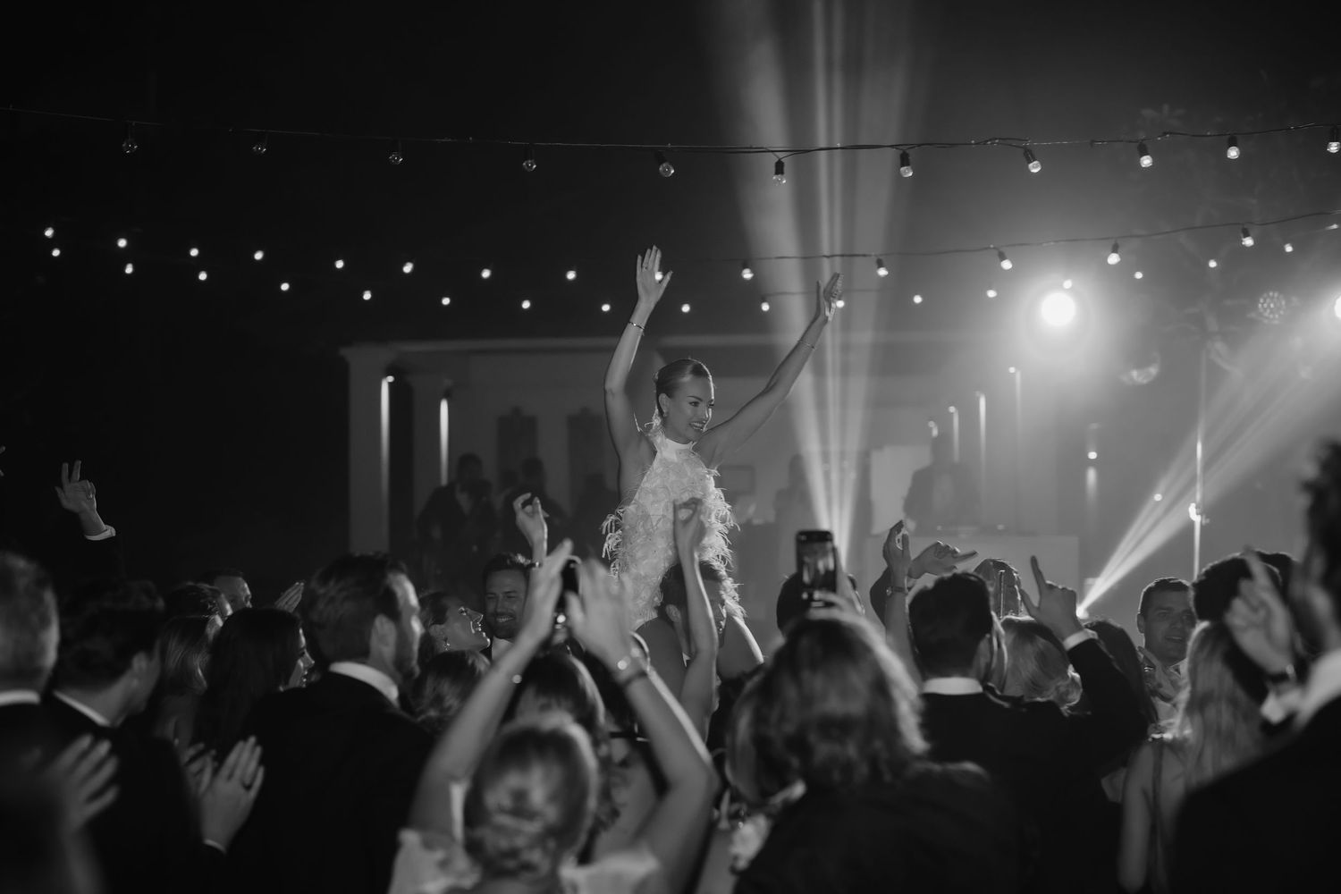Dramatic black and white shot of wedding celebration with string lights and spotlights illuminating the dance floor.