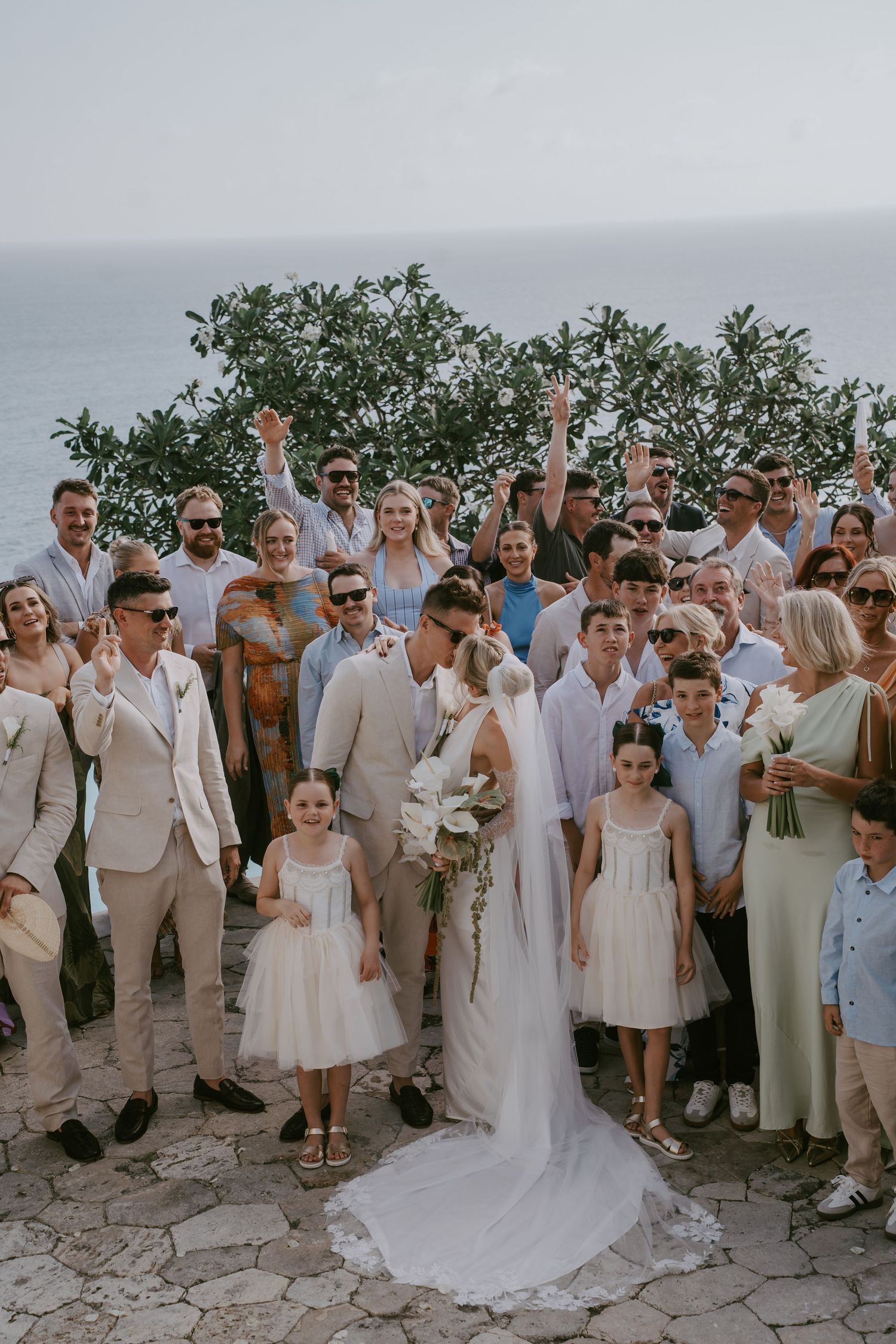 Large wedding party group photo with guests in light colored attire on a stone terrace.