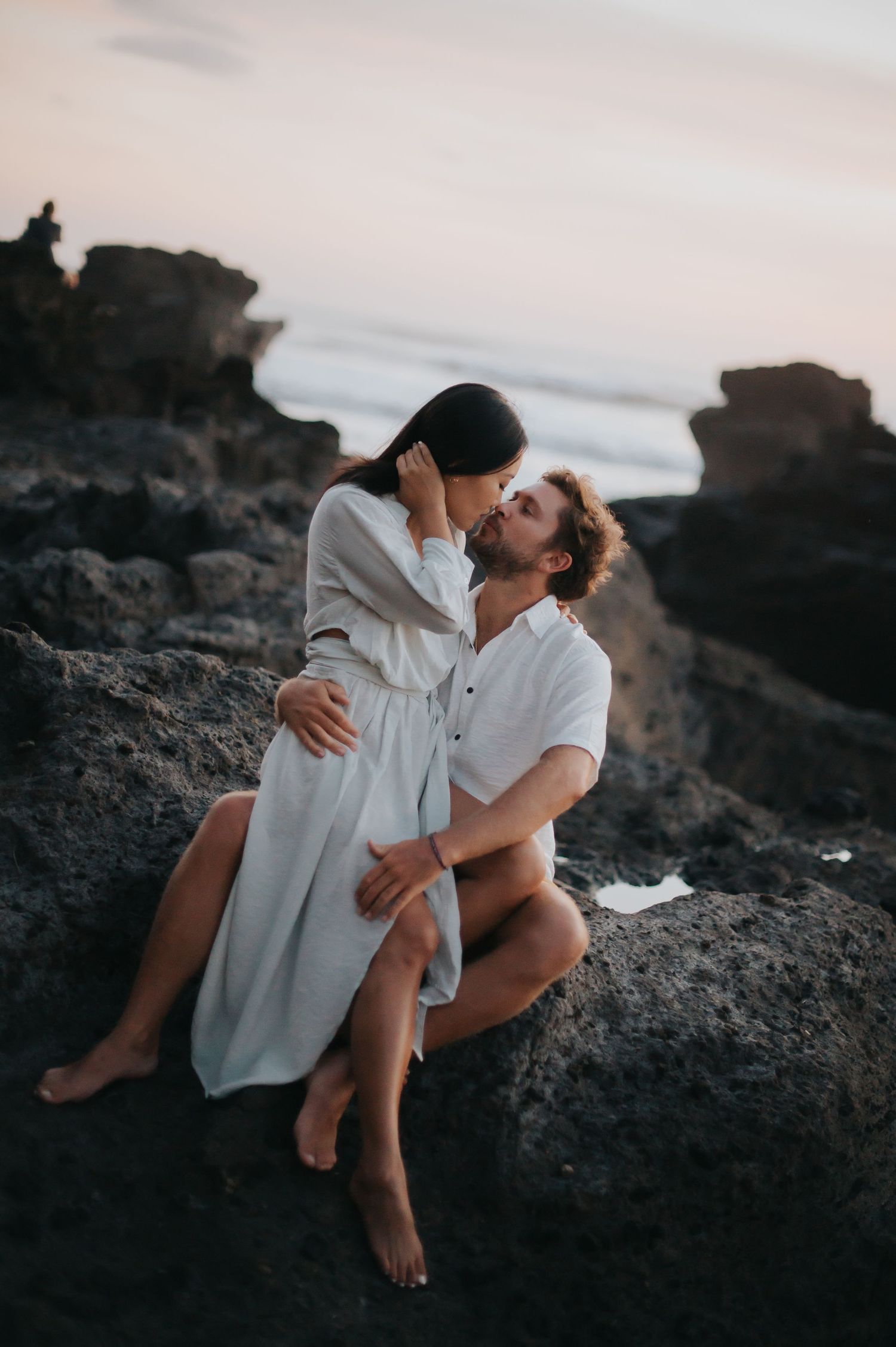 A romantic couple embraces on dark volcanic rocks at sunset in matching white outfits.
