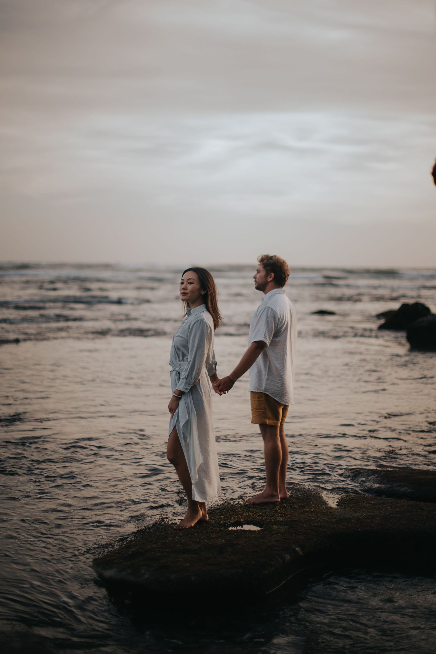 Two people stand silhouetted on rocky coastline at sunset holding hands as waves crash against the shore.