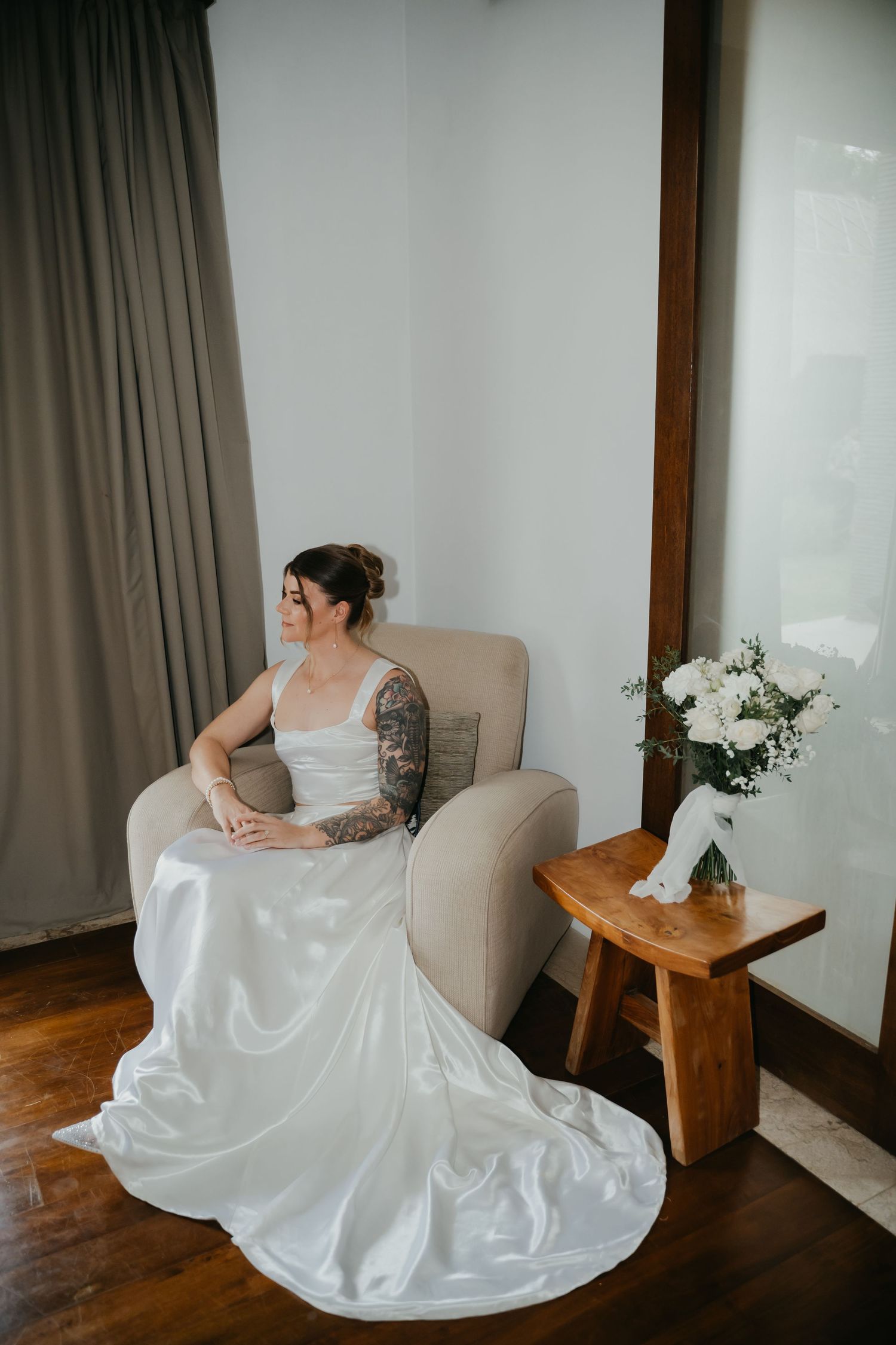 Tattooed bride in satin wedding dress with long train sits gracefully in armchair beside white floral bouquet.