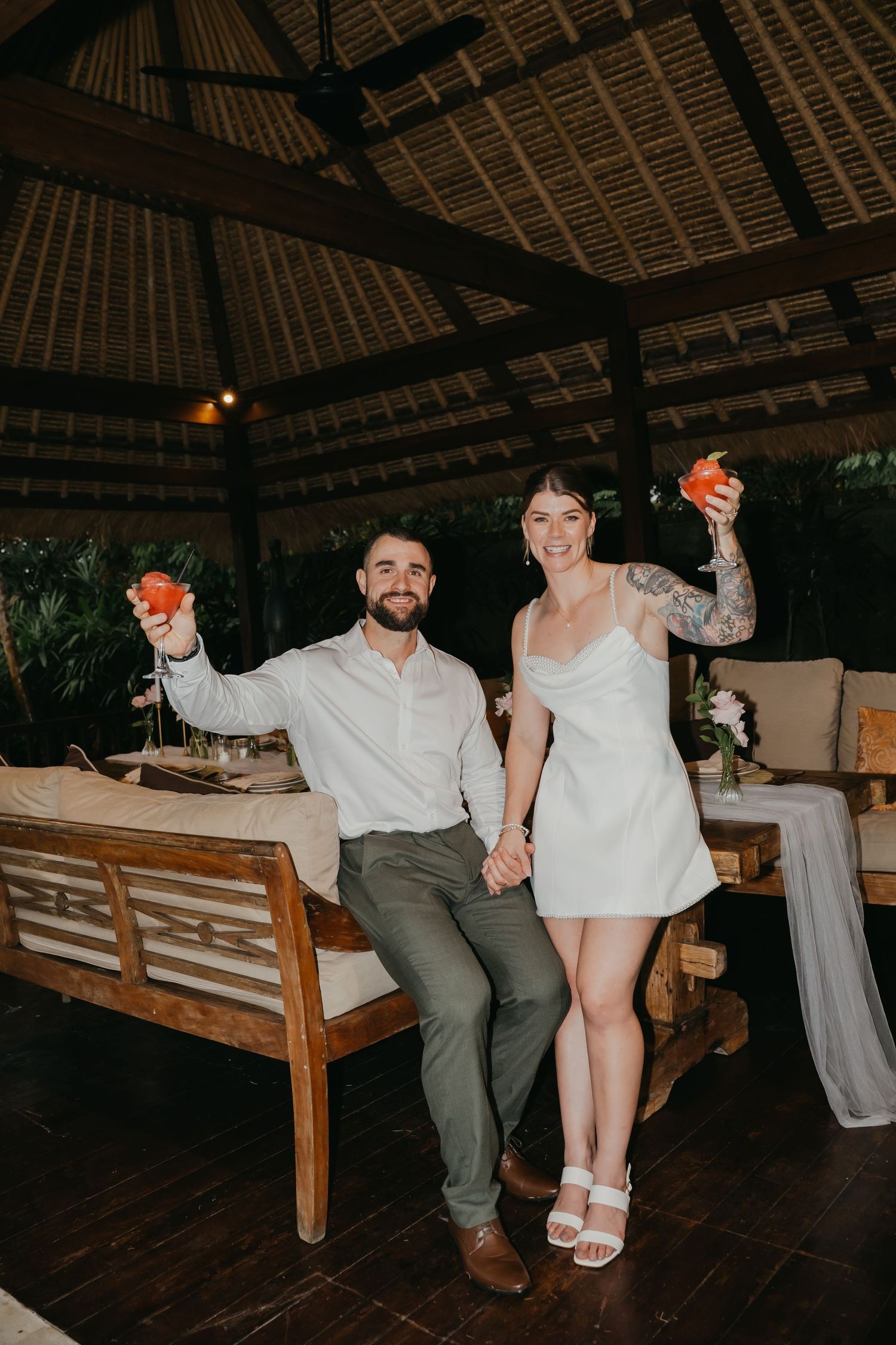 Couple celebrating at tropical reception venue, raising colorful cocktails in Balinese open-air pavilion.