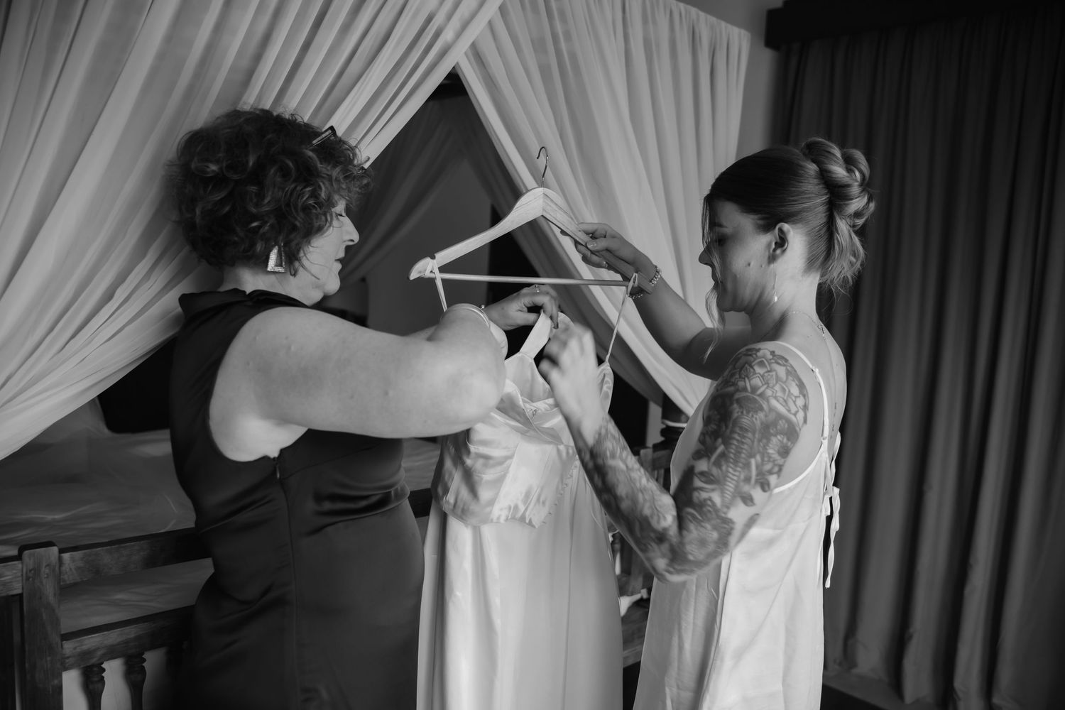 Black and white photo of two women handling a wedding dress on a hanger indoors.