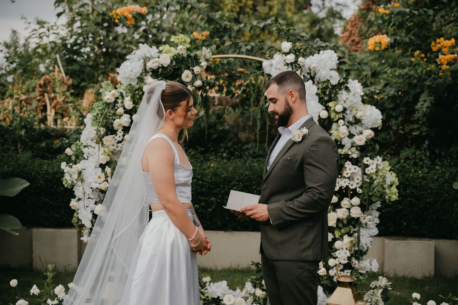 Bride and groom exchanging vows under white floral arch in lush garden wedding ceremony.