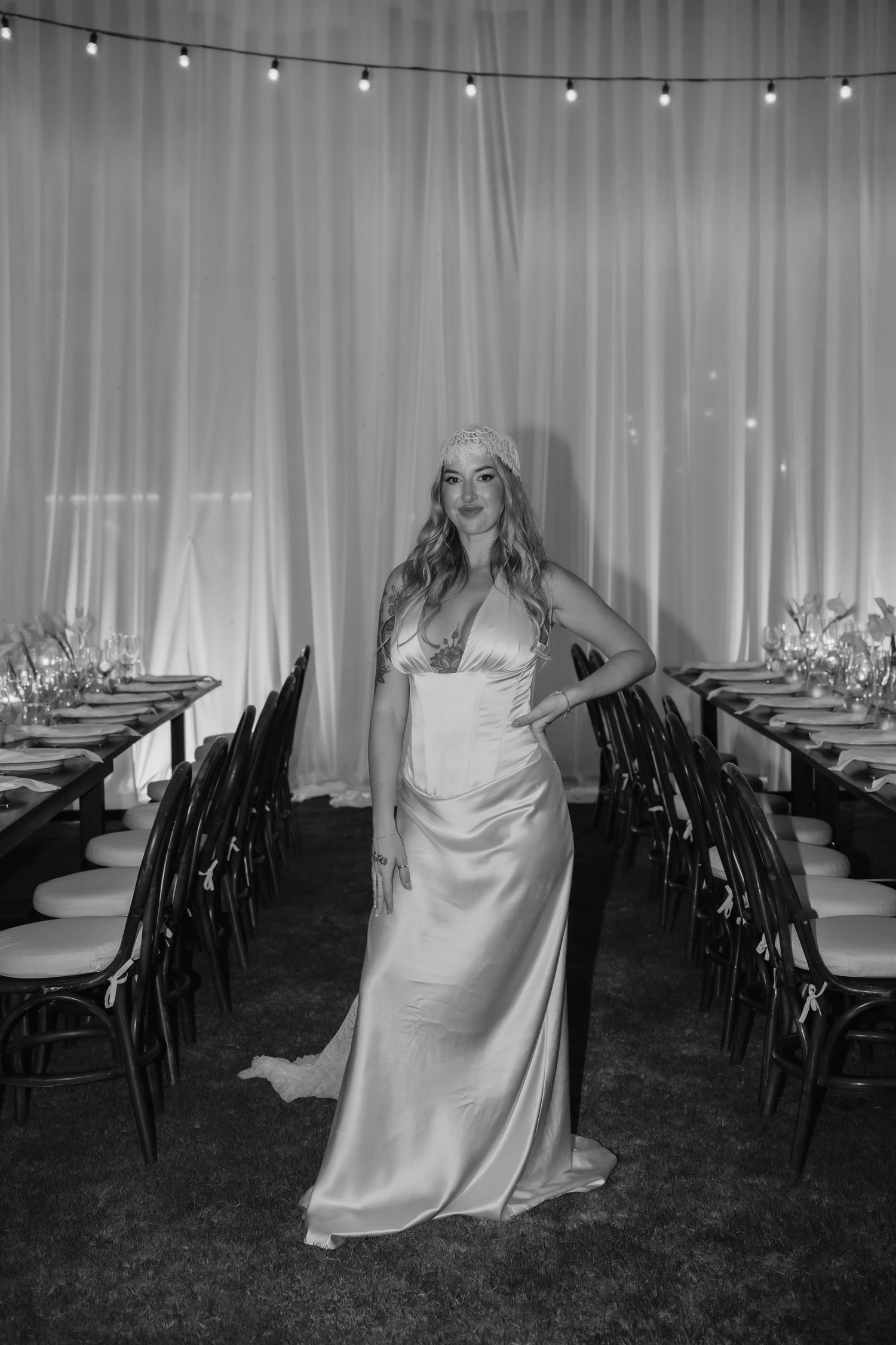 Black and white portrait of bride in satin gown and lace headpiece standing in decorated reception venue.