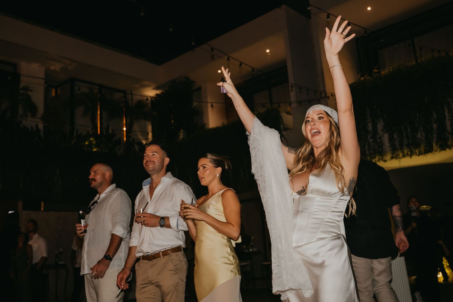 Bride celebrates joyfully with raised arms and sparkler surrounded by guests at evening wedding reception.