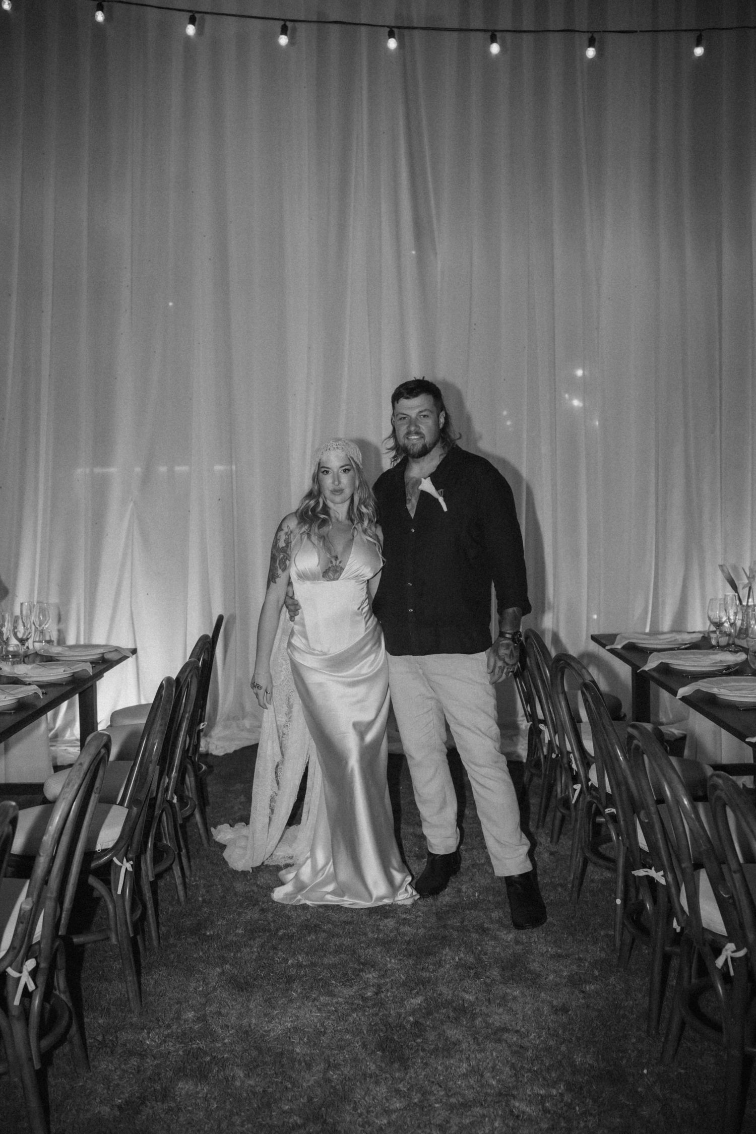 Black and white photo of a bride and groom standing together in a decorated wedding reception venue with string lights.