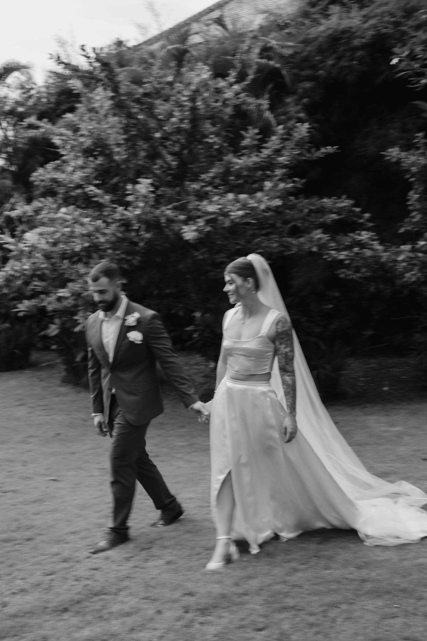 Black and white photo of a bride and groom holding hands, walking outdoors with lush greenery behind them.