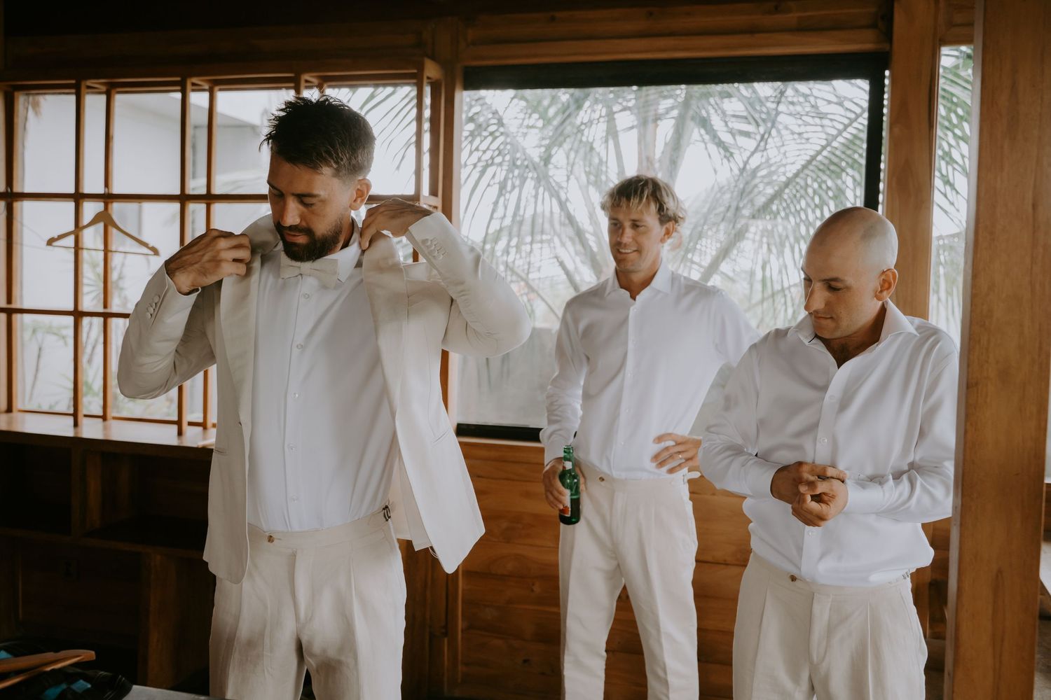 Groom and groomsmen dressed in white linen suits preparing for wedding ceremony in tropical wooden venue.