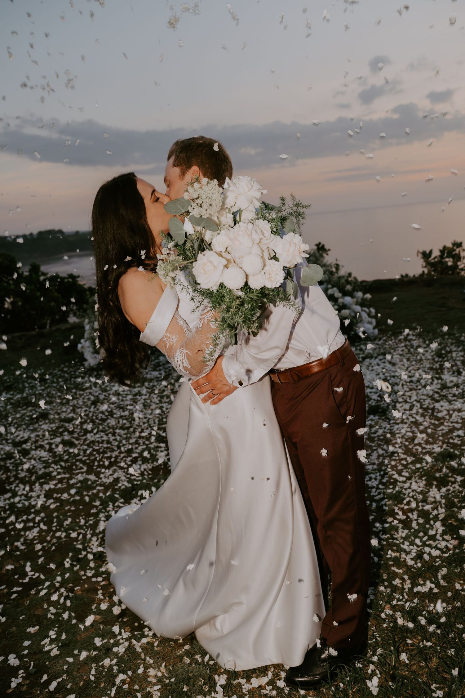 Bride and groom share a first kiss surrounded by falling white flower confetti petals during their outdoor sunset wedding ceremony.