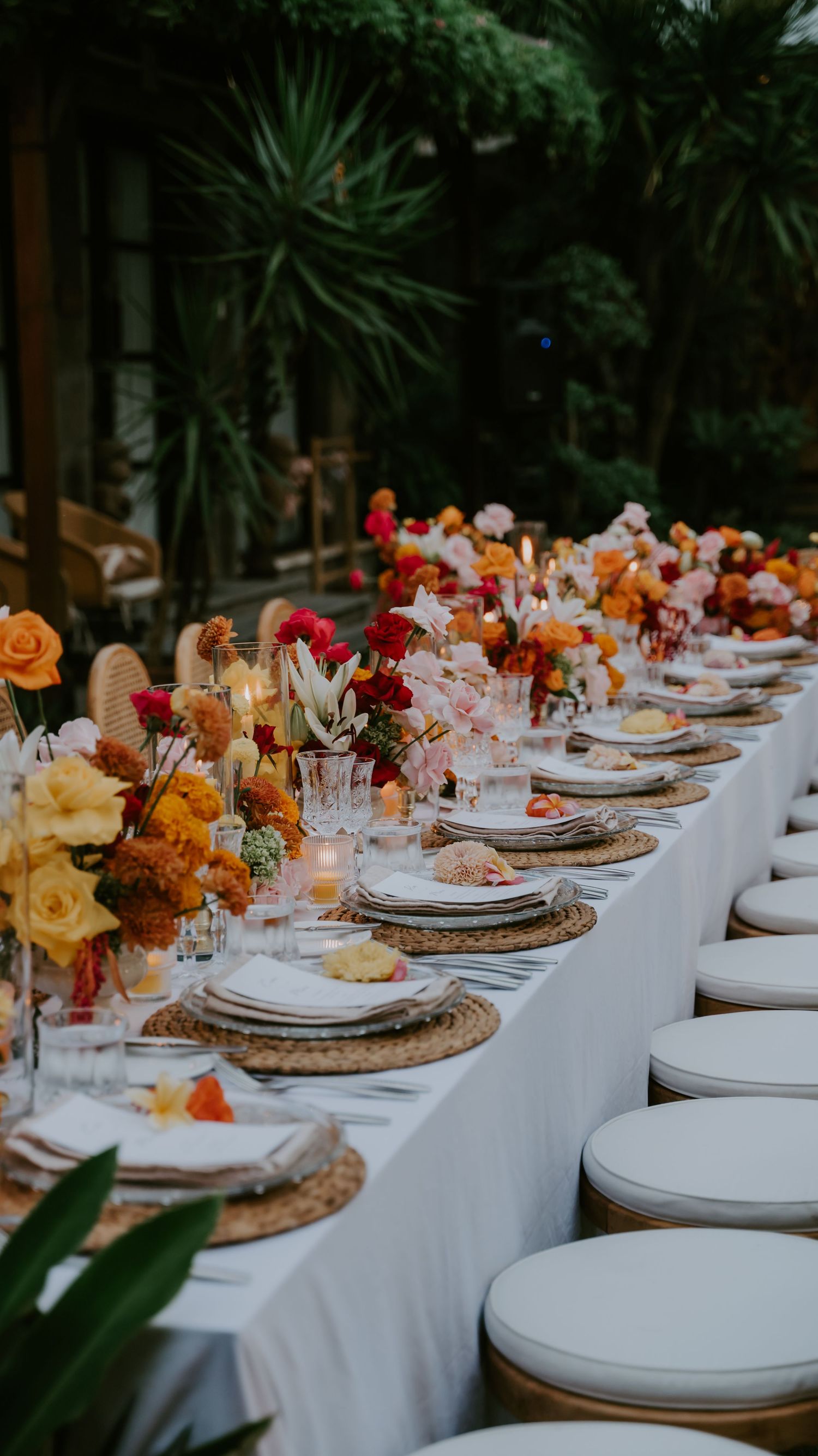 Luxurious outdoor wedding reception long table with vibrant orange and red floral centerpieces and white place settings.